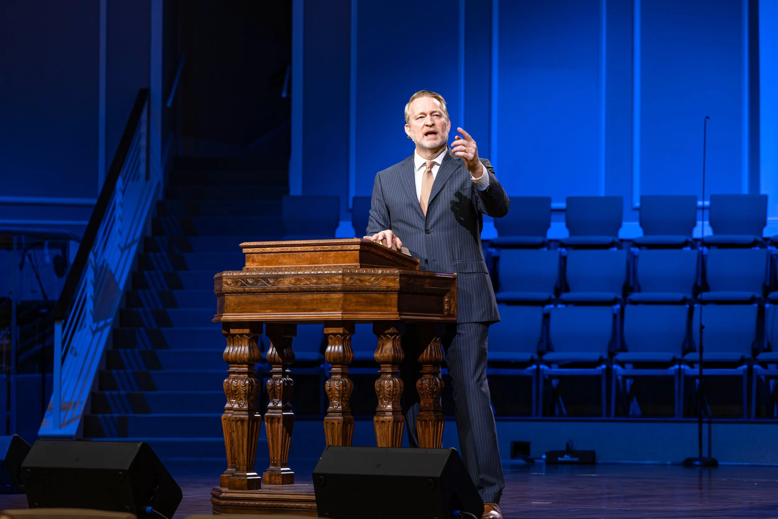 A man in a dark pinstripe suit and light-colored tie is speaking at a wooden podium on a stage with blue lighting. There are empty blue chairs in the background and a staircase on the left side of the stage.