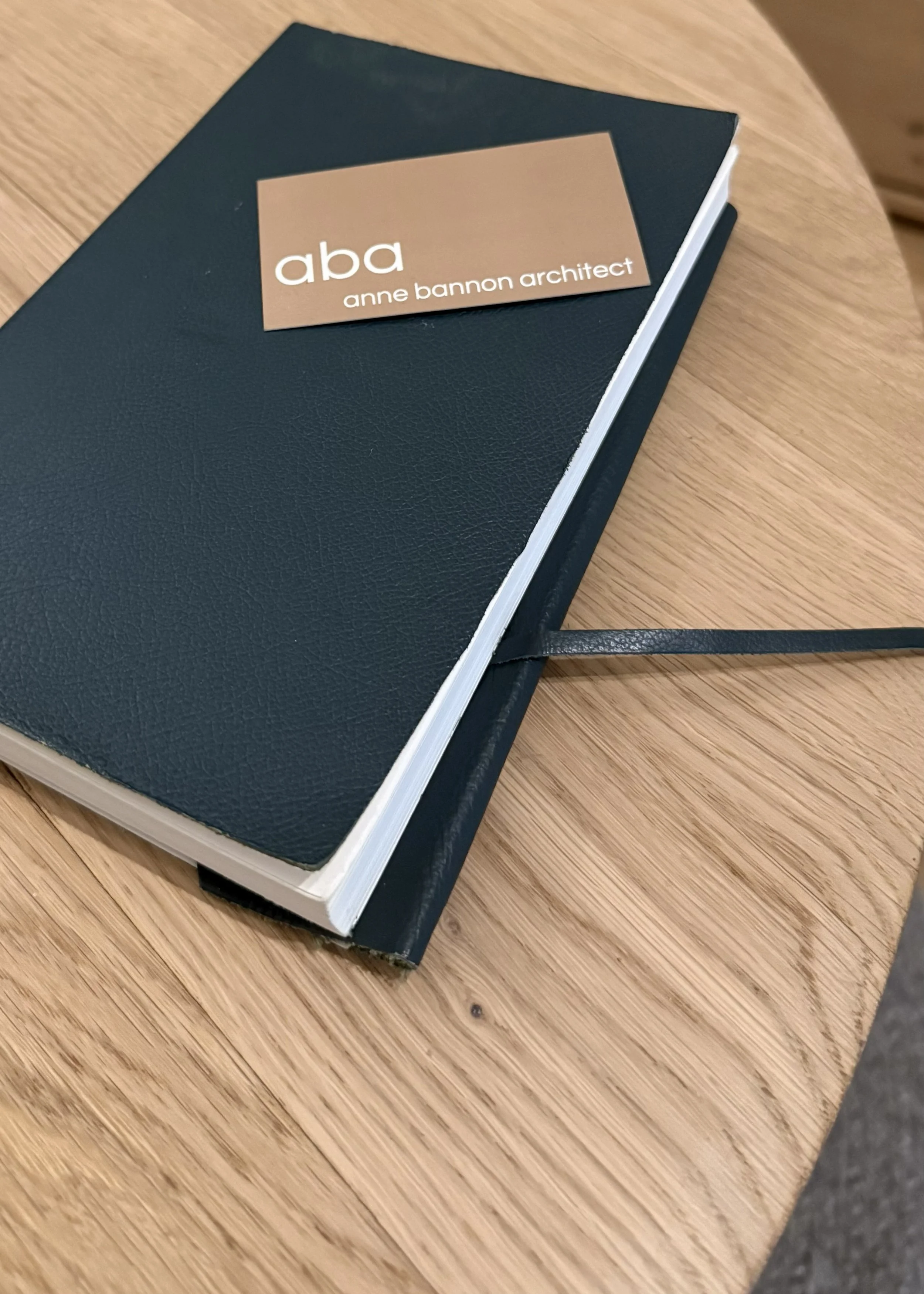 Close-up of a sketchbook with a business card that reads 'aba anne bannon architect' placed on a wooden surface.