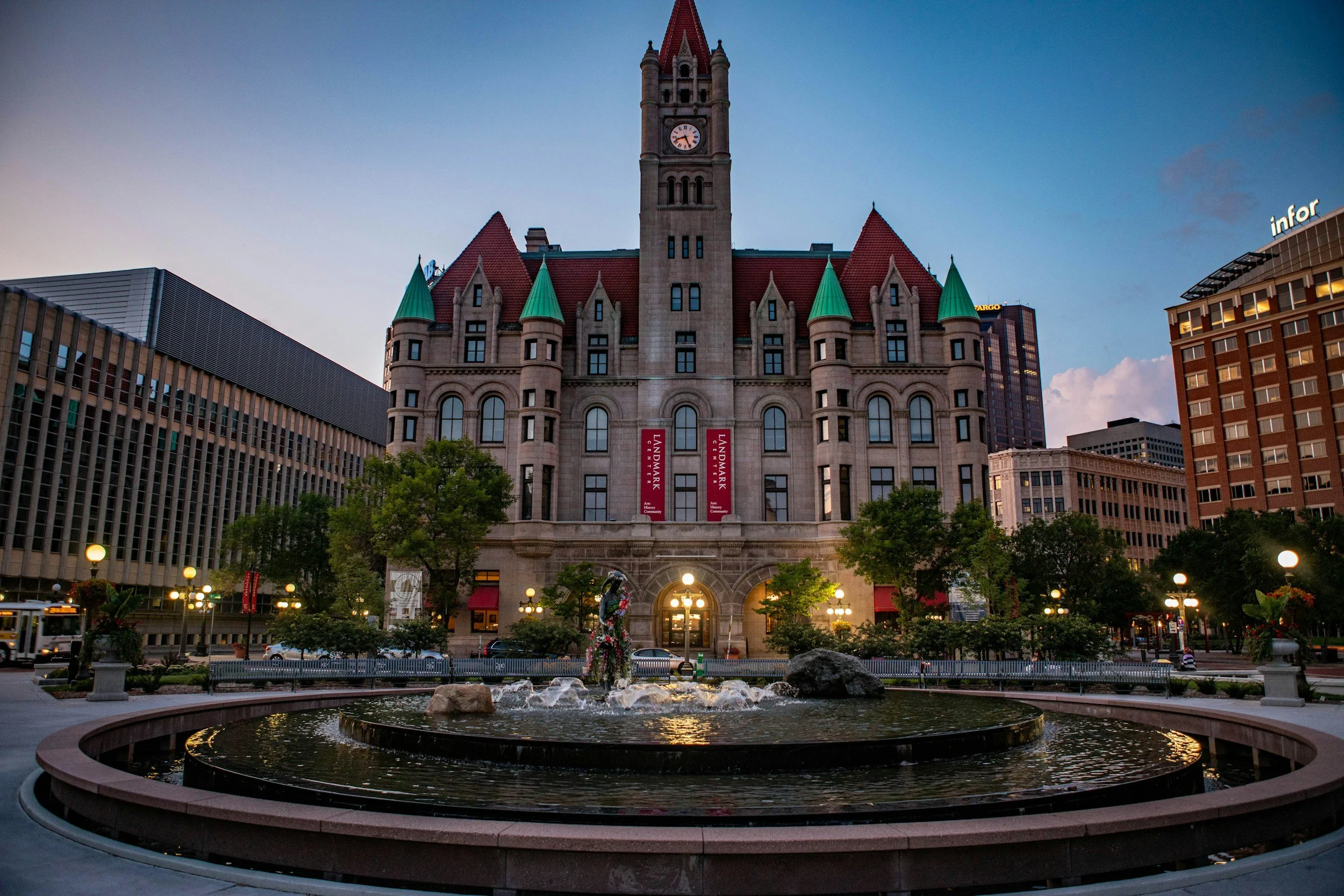 Historic Landmark Center with clock tower and red banners, trees and fountain in foreground, surrounding modern buildings, in an urban park setting at dusk.