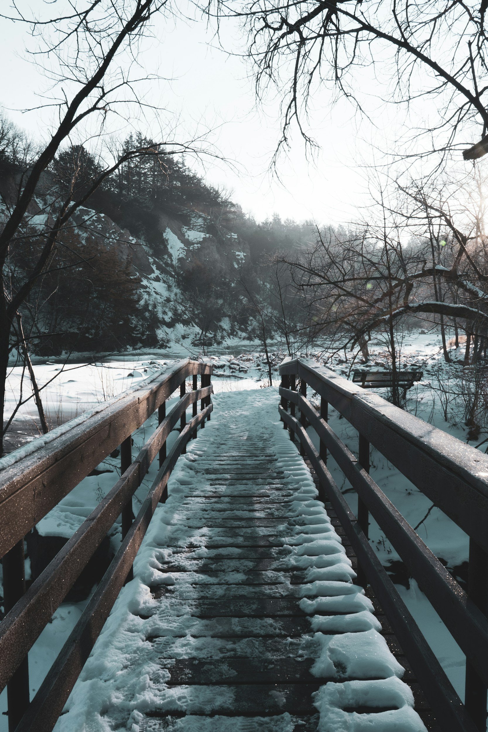 Snow-covered wooden bridge over a river in winter, with leafless trees and mountains in the background.