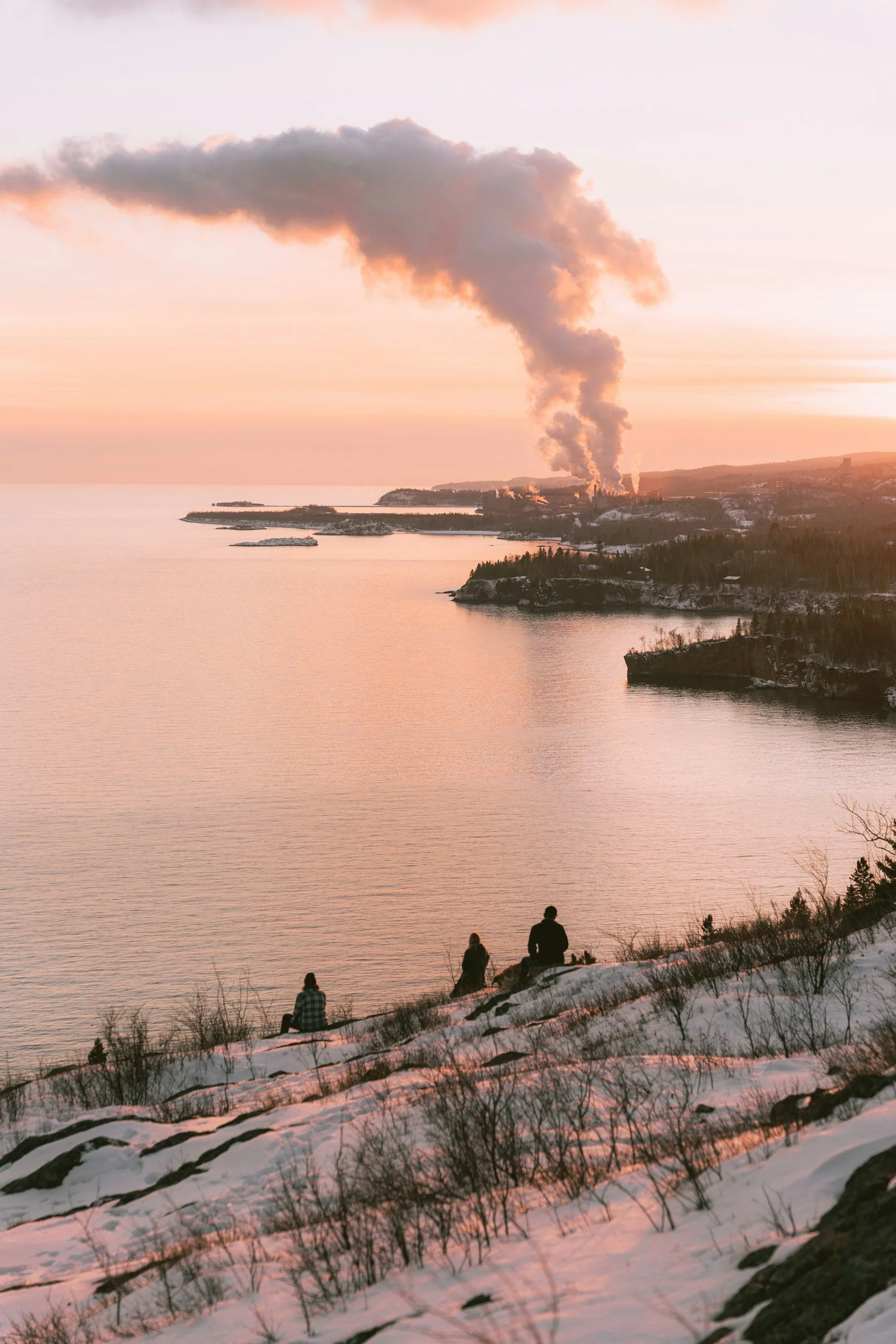 People sitting on a snowy hillside watching a sunset over a body of water with industrial smoke plumes in the distance.