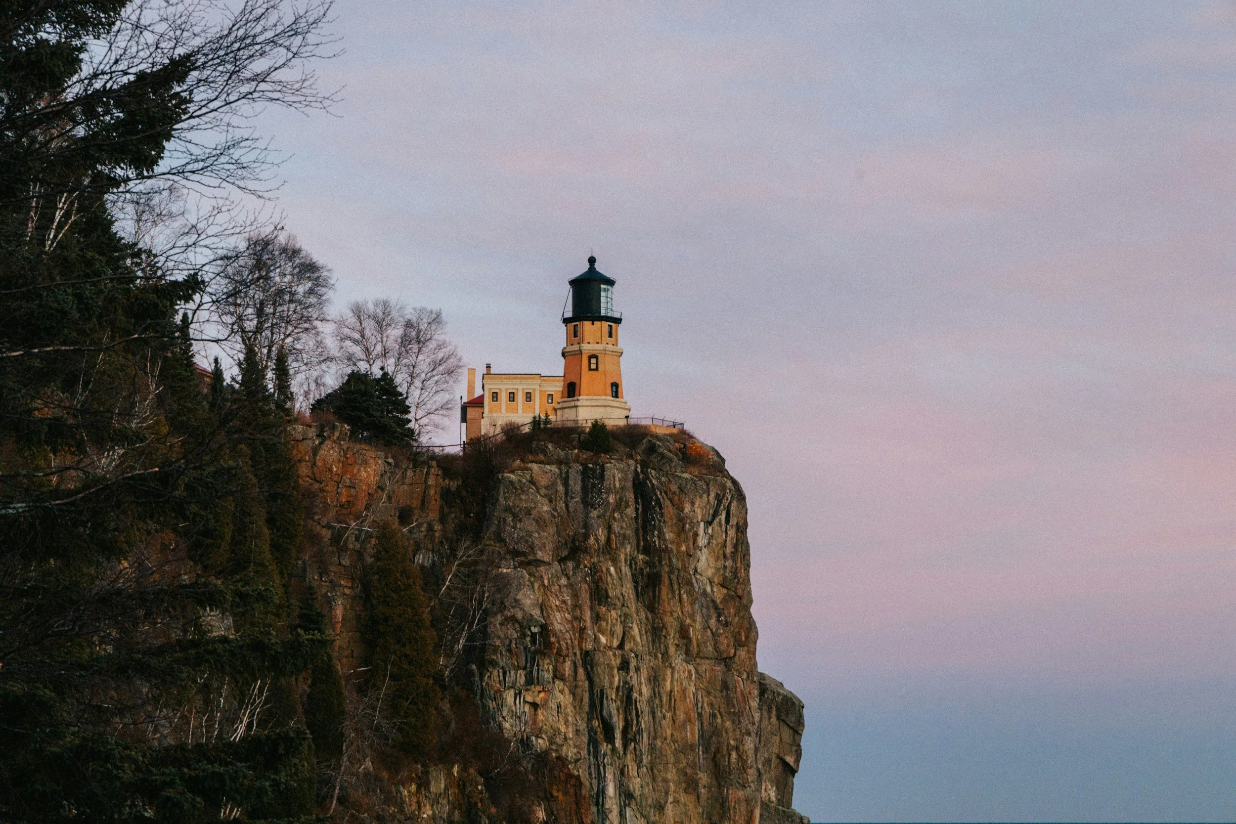 Split Rock Lighthouse situated on the edge of a rocky cliff with trees on the left, under a pastel-colored sky.