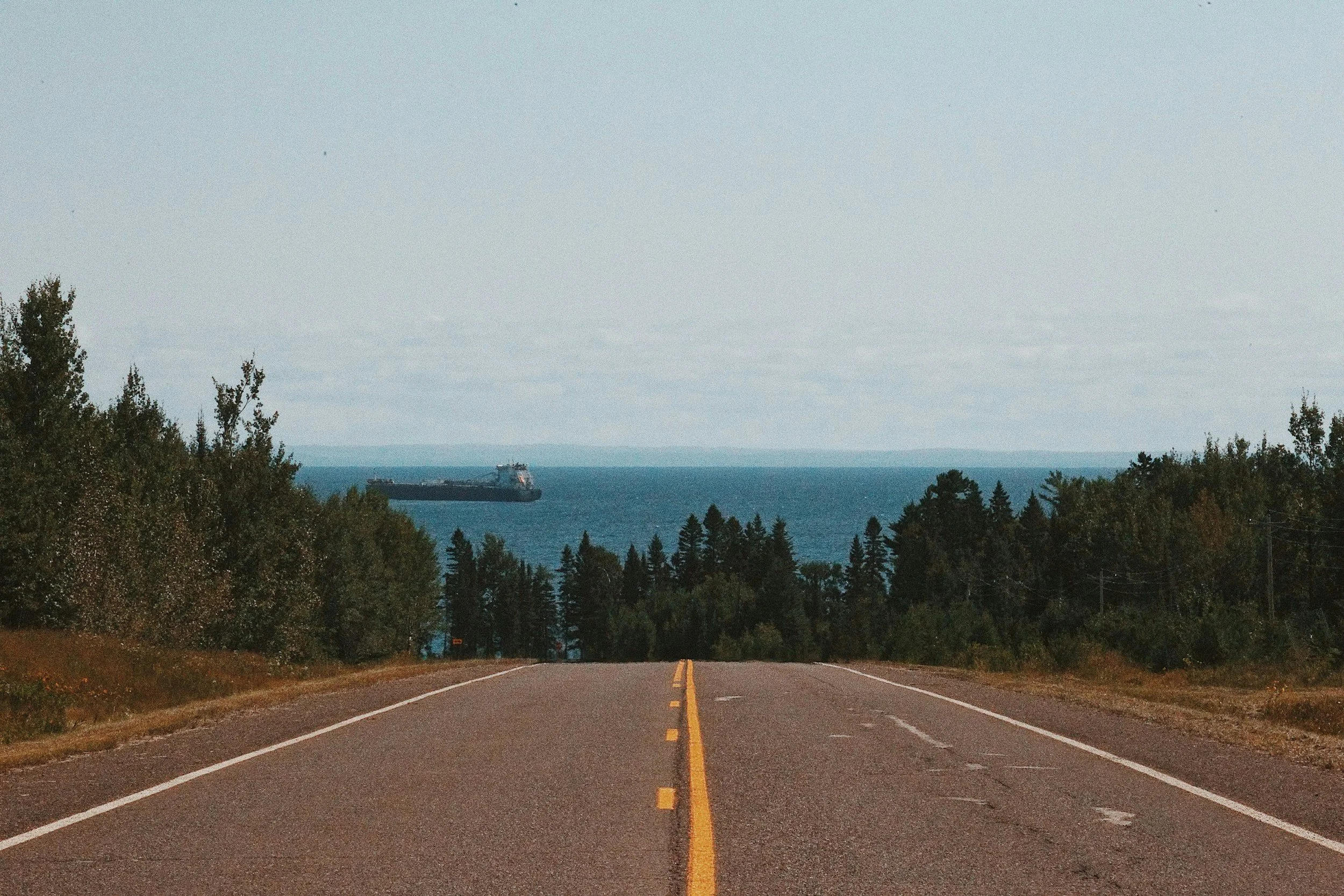 A two-lane road leading towards Lake Superior with a cargo ship in the distance, surrounded by trees on both sides.