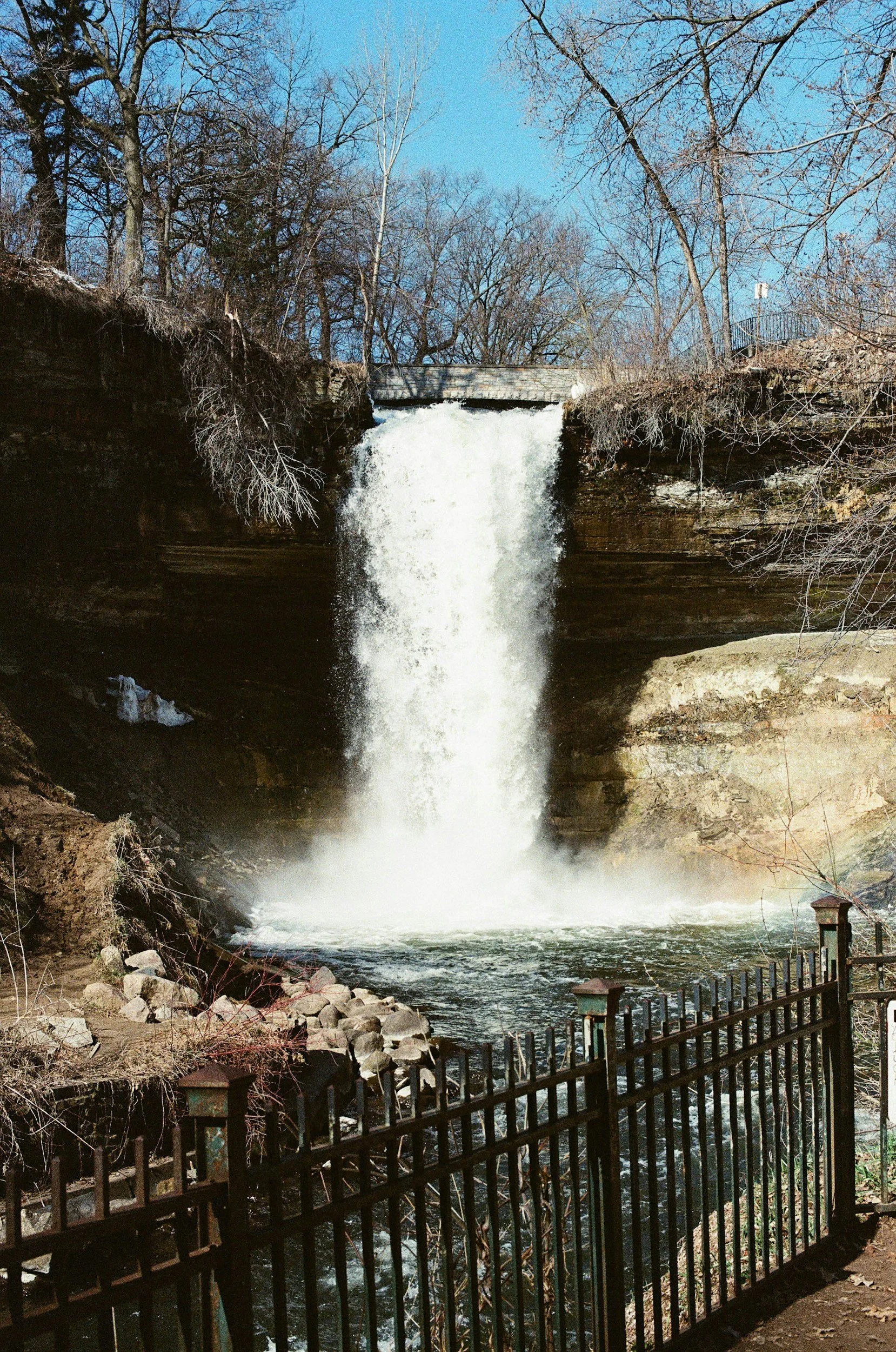 Minnehaha Falls flowing over a cliff into a pool below, surrounded by leafless trees and a black metal fence in the foreground.