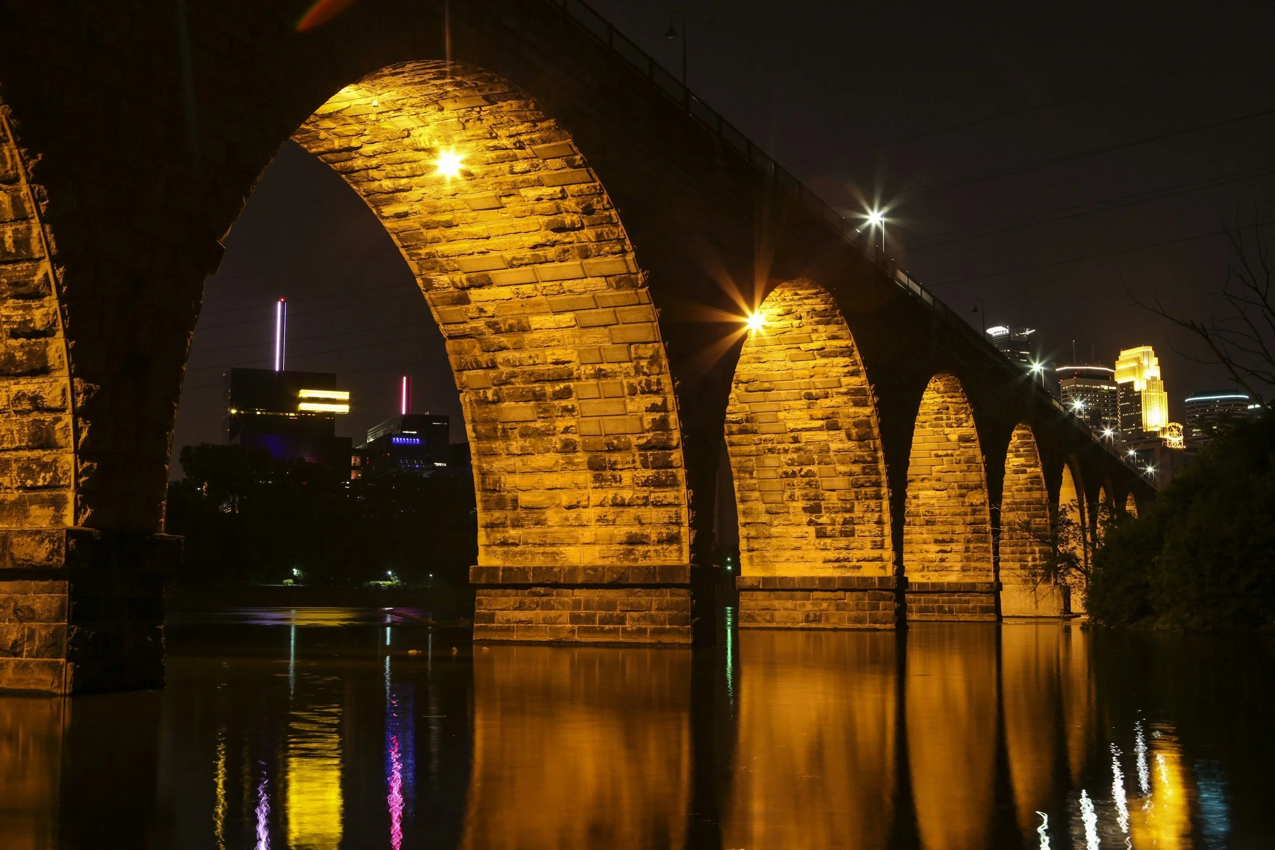 Night view of a historic Stone Arch Bridge with multiple arches illuminated by yellow lights, reflecting on the water below, with a city skyline with tall buildings and illuminated signs in the background.