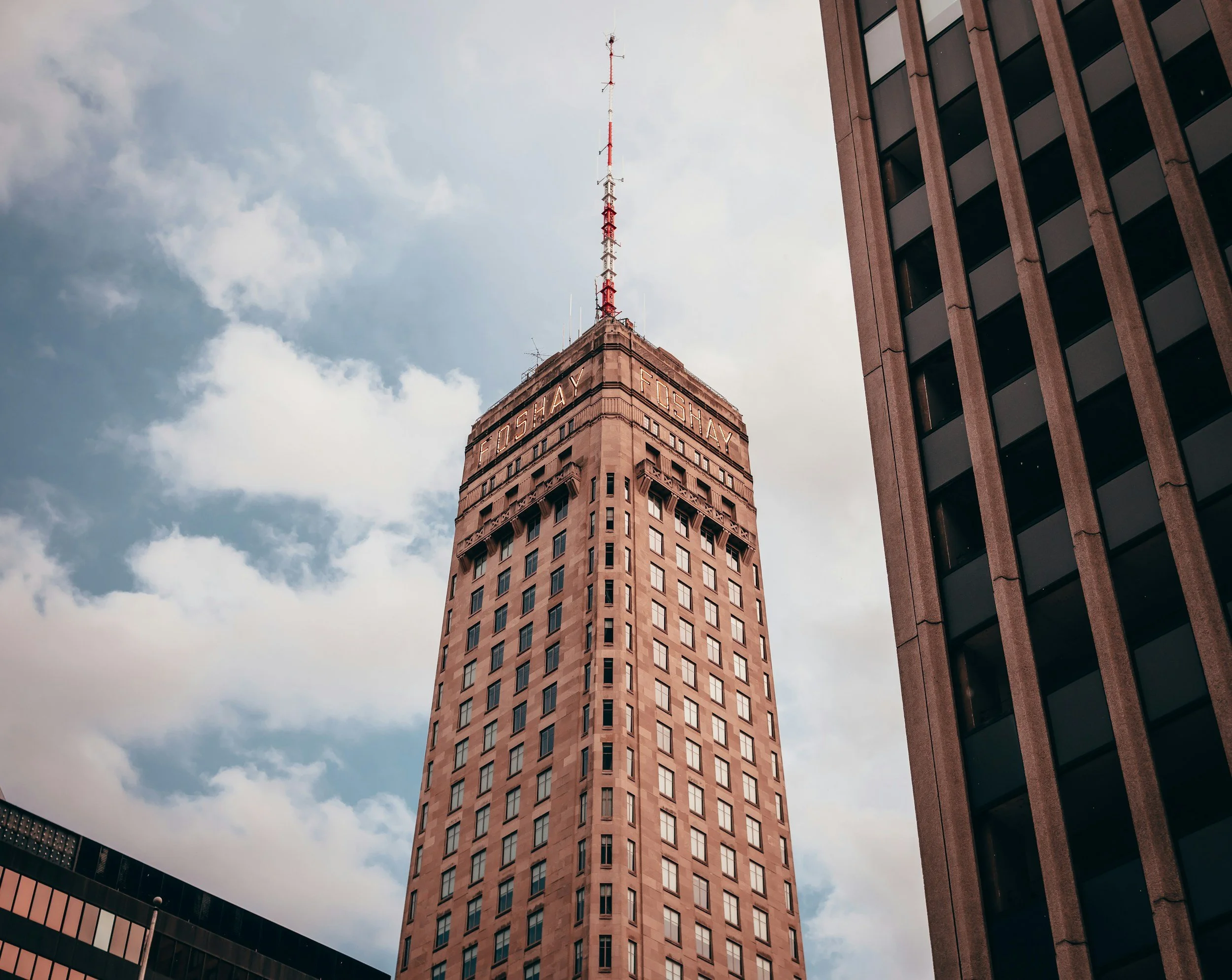 An upward shot of the Foshay Tower skyscraper with a tower antenna on top, surrounded by other tall buildings, under a partly cloudy sky.