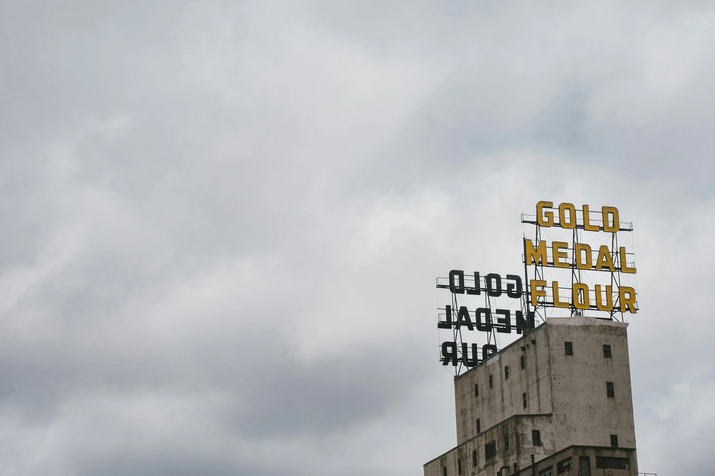 Sign on top of a building reads "GOLD MEDAL FLOUR" with some letters flipped or missing, against a cloudy sky.