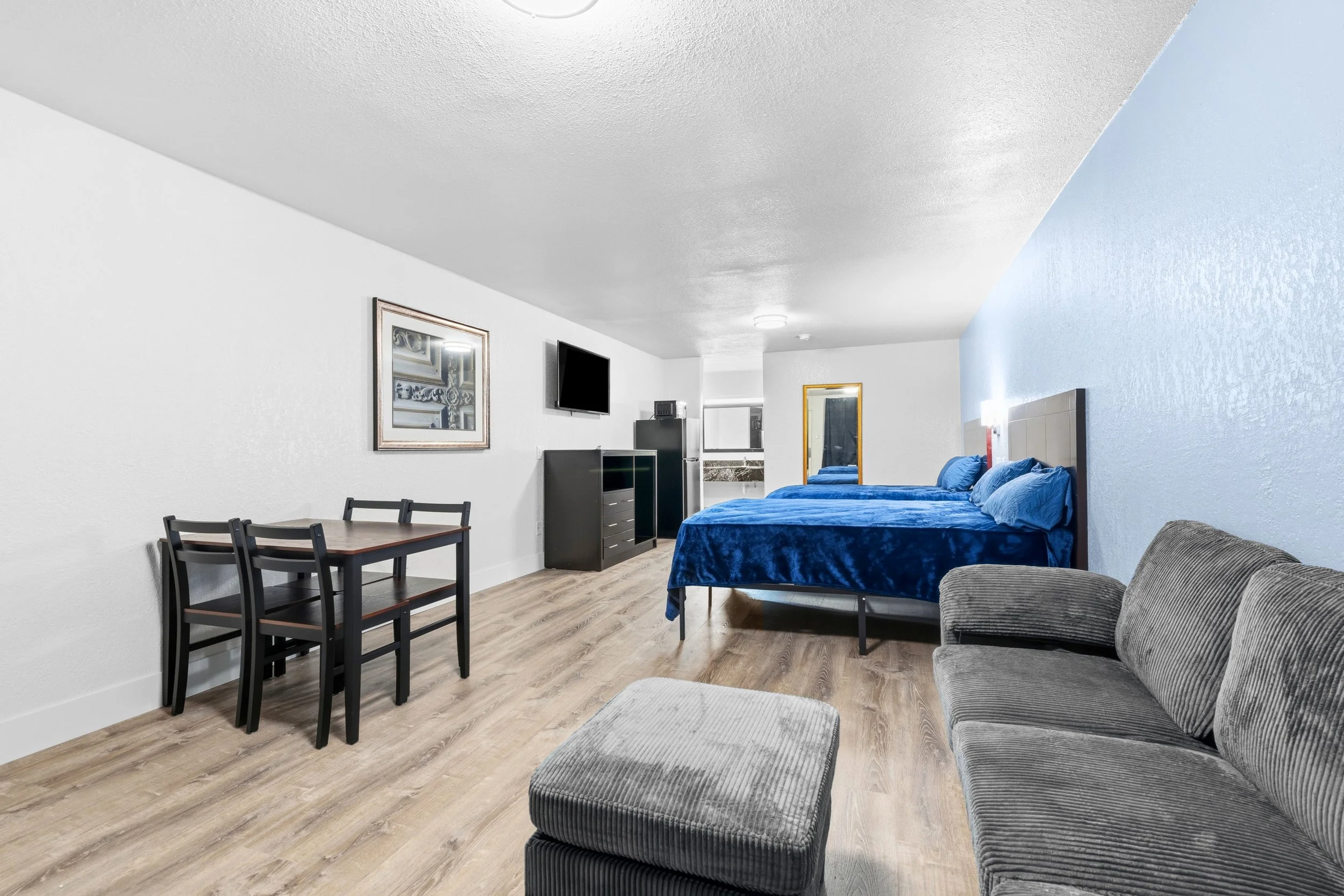 A hotel room with a double bed with a blue blanket, a gray sofa with a matching footstool, a small dining table with four chairs, a wall-mounted TV, and a mirror on the wall.