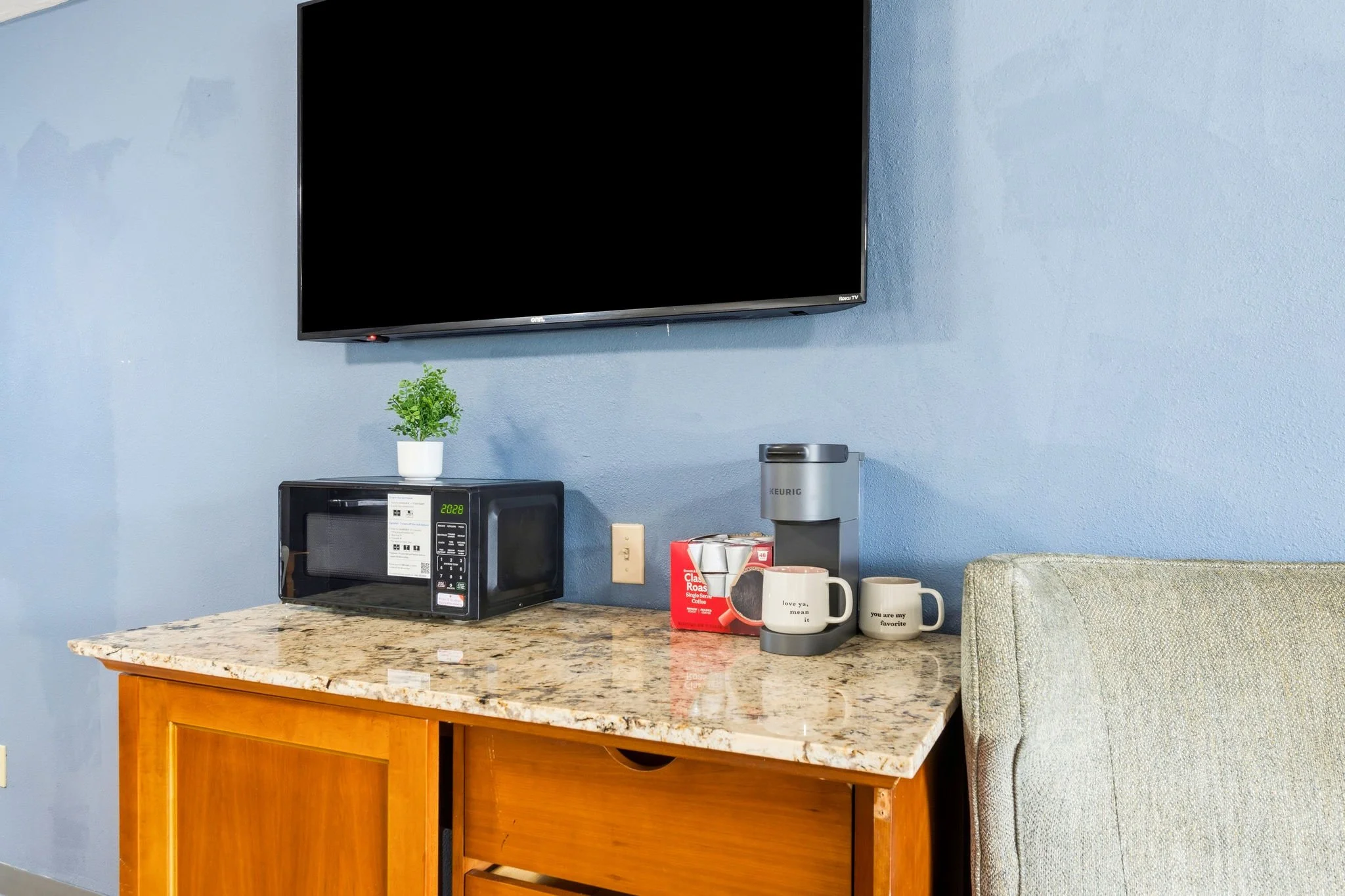 A kitchen countertop with a microwave oven, a potted plant, a red coffee packet, two mugs, and a Keurig coffee maker against a blue wall with a wall-mounted TV.