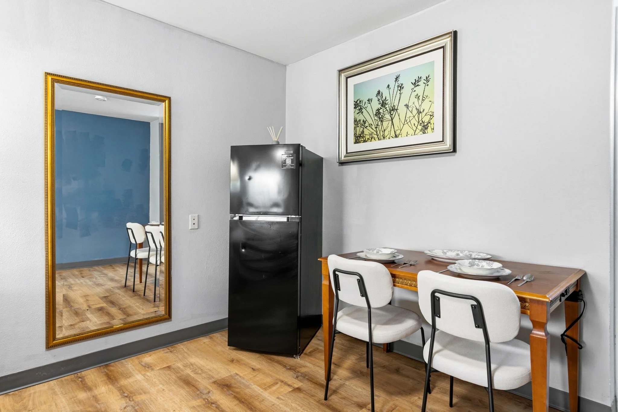 A dining room with a wooden table set with white and blue patterned plates, placed with silverware. Four white chairs with black frames surround the table. A black water cooler stands against a white wall. A framed picture of plant silhouettes hangs 