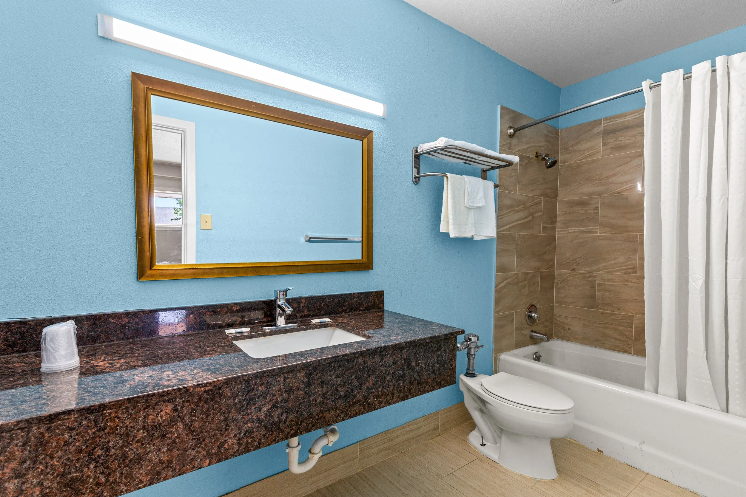 A bathroom with blue walls, a granite countertop with a sink, a mirror with a wooden frame, towels on a metal rack, a bathtub with a shower curtain, and beige tile flooring.