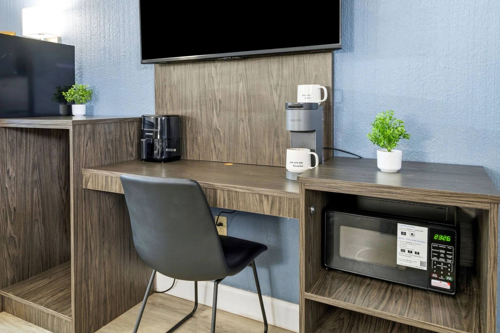 A small kitchenette area with a wall-mounted flat-screen TV, a wooden countertop, a coffee maker, a white mug, a small green potted plant, and a microwave oven on a lower shelf against a blue wall.