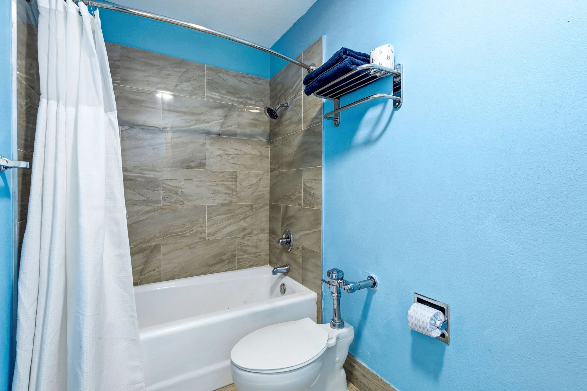 Bathroom with blue wall, bathtub, showerhead, tiled wall, white shower curtain, toilet, and towel rack with dark towels and toilet paper.