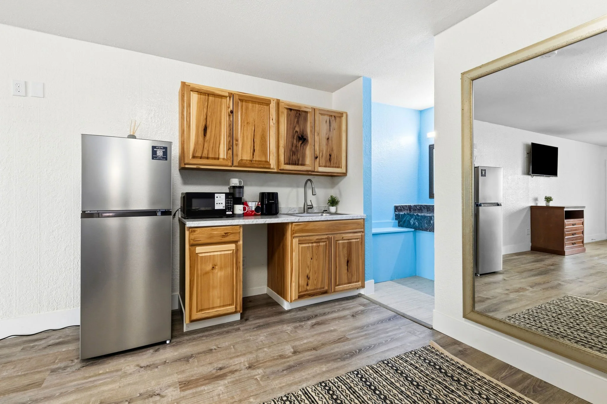 Kitchen with wooden cabinets, stainless steel refrigerator, microwave, coffee maker, and a small potted plant, with a doorway leading to a blue-painted room.