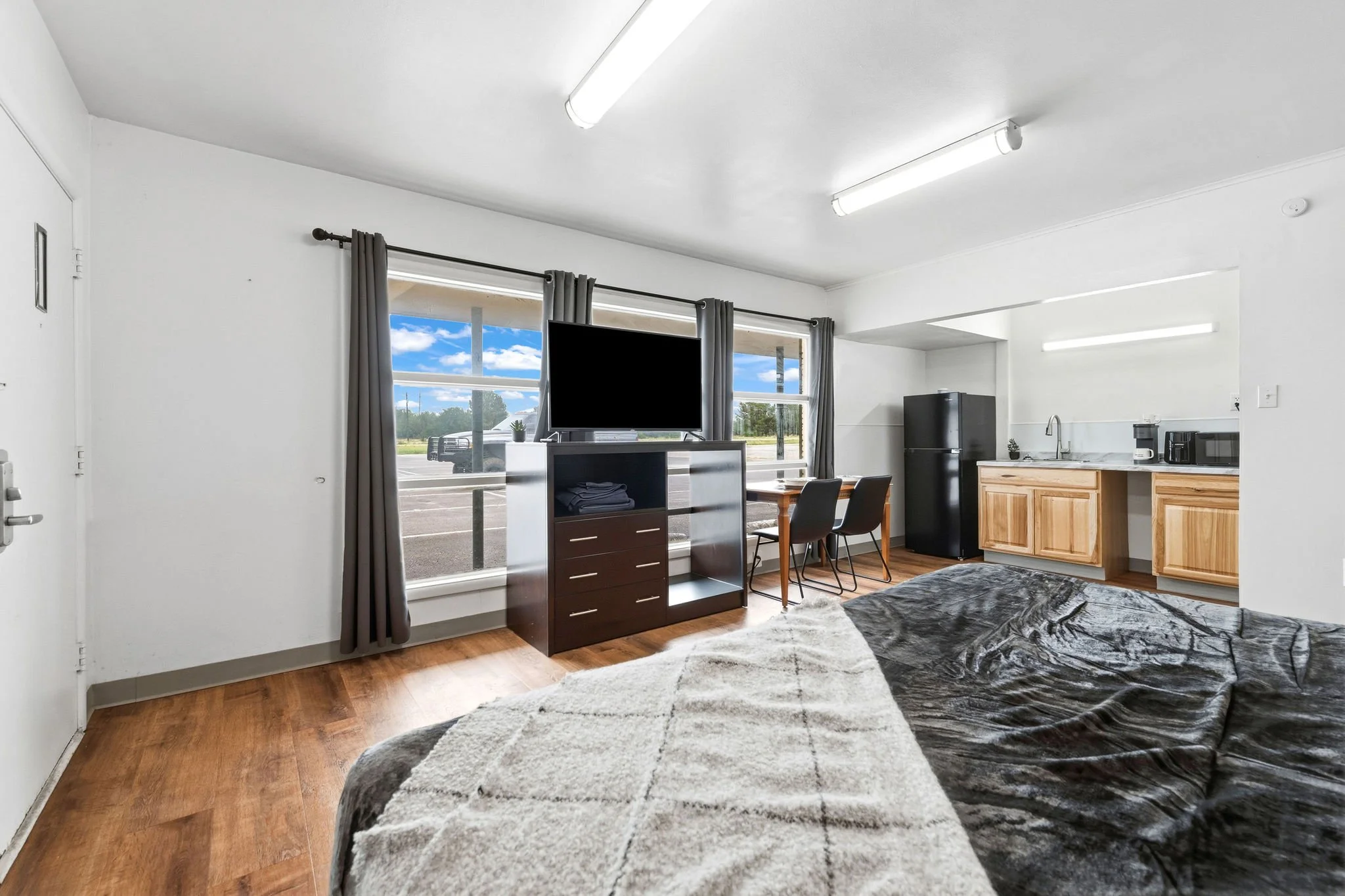 Interior of a small apartment with a bed, a TV on a stand, a kitchen area with a mini fridge and small appliances, and windows showing a blue sky with clouds.