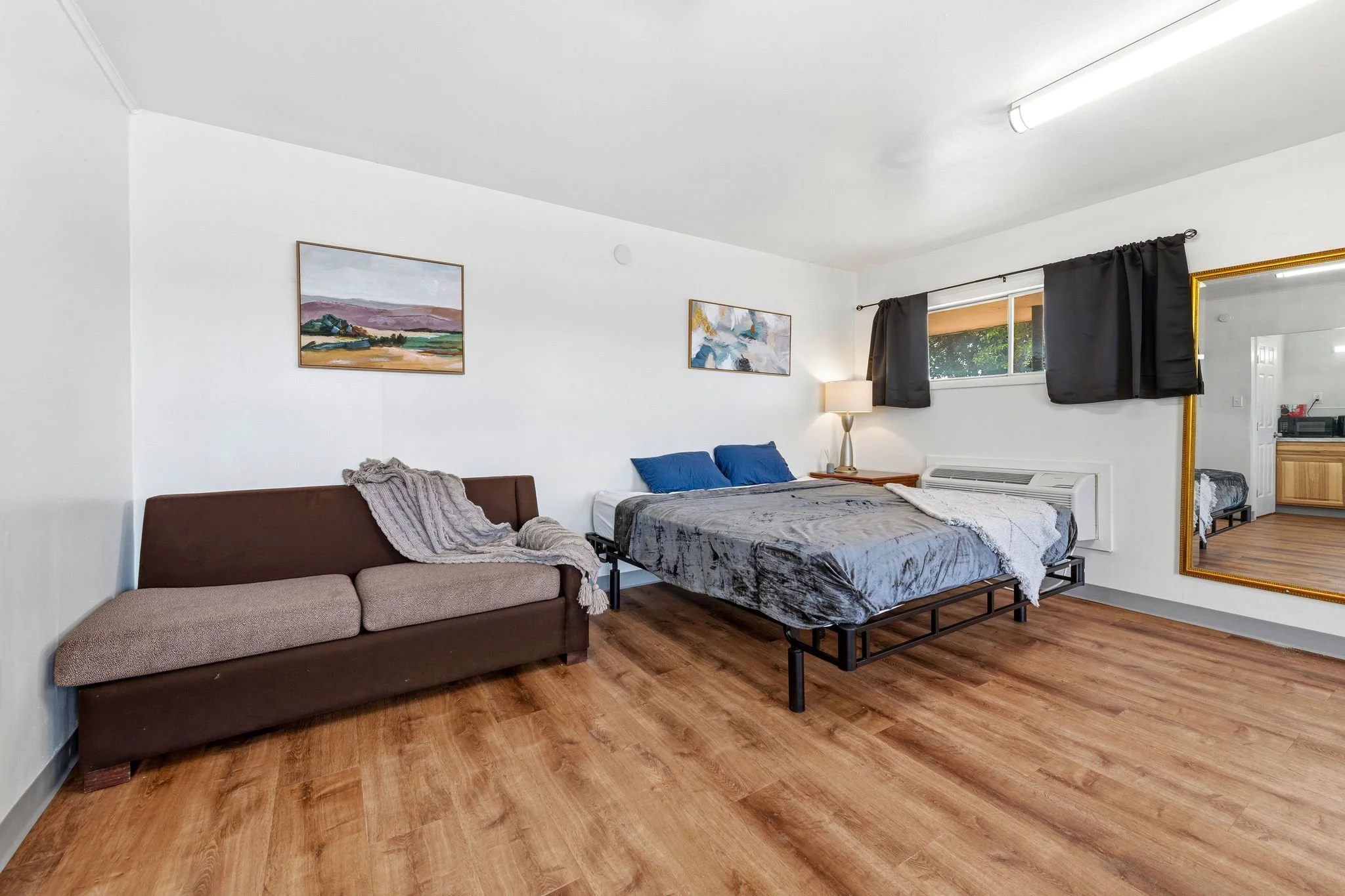 A furnished bedroom with wooden flooring, a brown sofa with a blanket, a bed with blue pillows, a nightstand with a lamp, a large mirror with a gold frame, and wall art depicting landscape scenes.