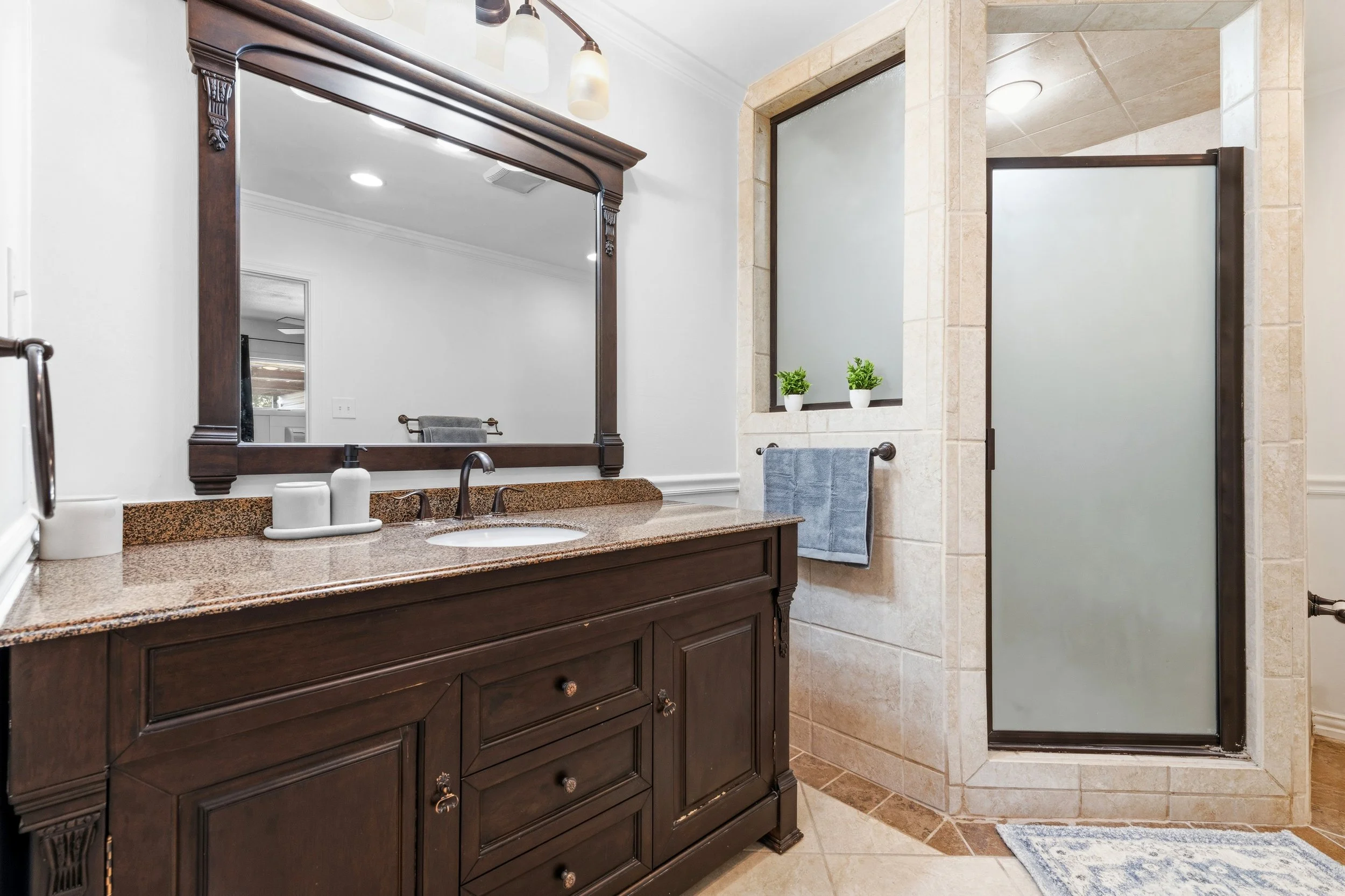 Bathroom with dark wood vanity, granite countertop, large mirror, frosted glass shower door, two small potted plants on a ledge, towel bar with blue towel, and a blue rug.