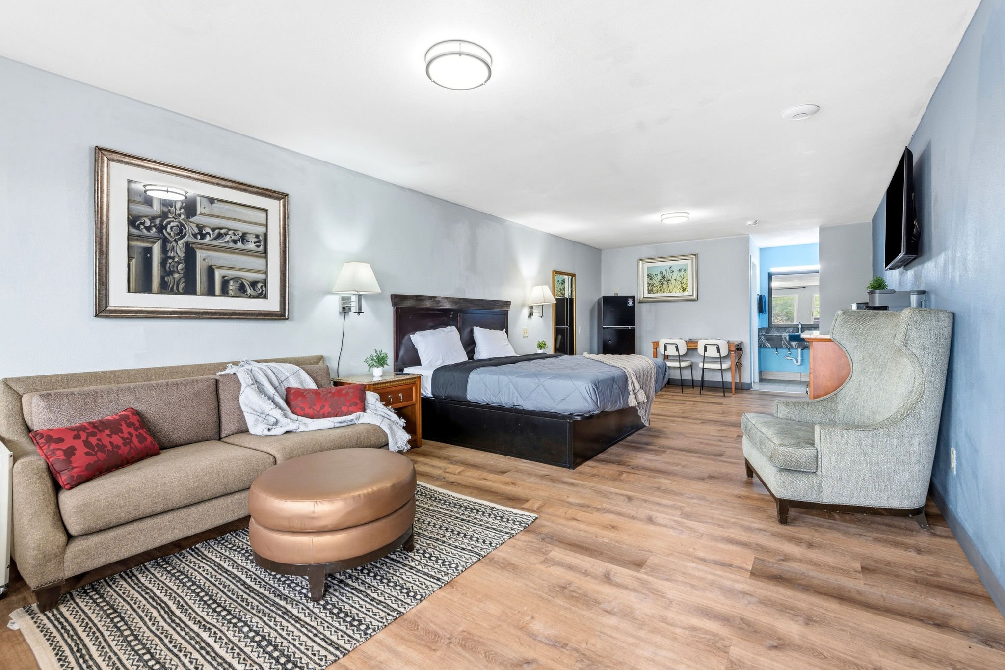 Interior view of a hotel room with a bed, sofa, armchair, and artwork on light blue walls, with wood flooring and ceiling lights.