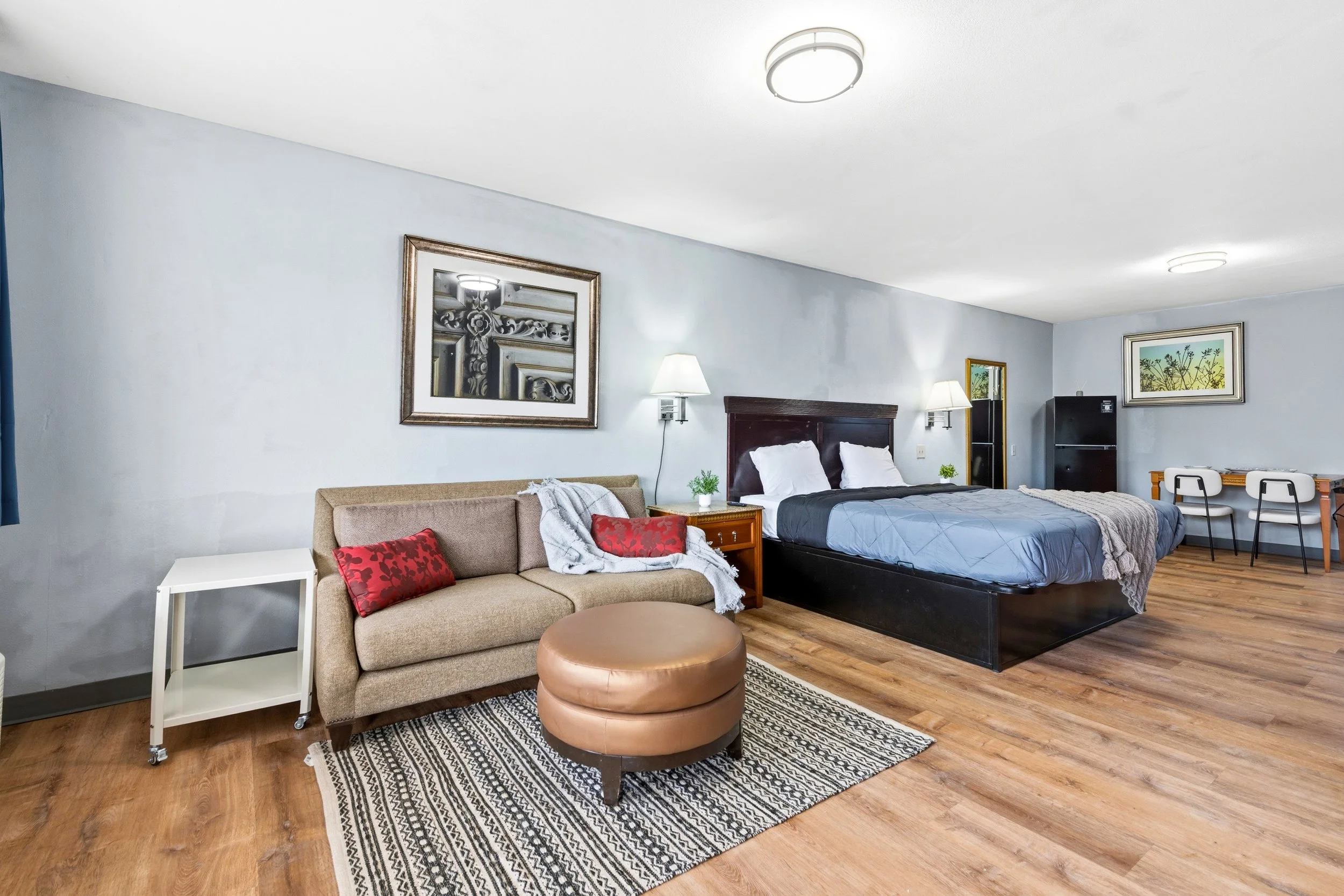 Interior of a hotel room with a beige sofa with red cushions, a round ottoman, a bed with white pillows, lamps, framed artwork, and a small table.