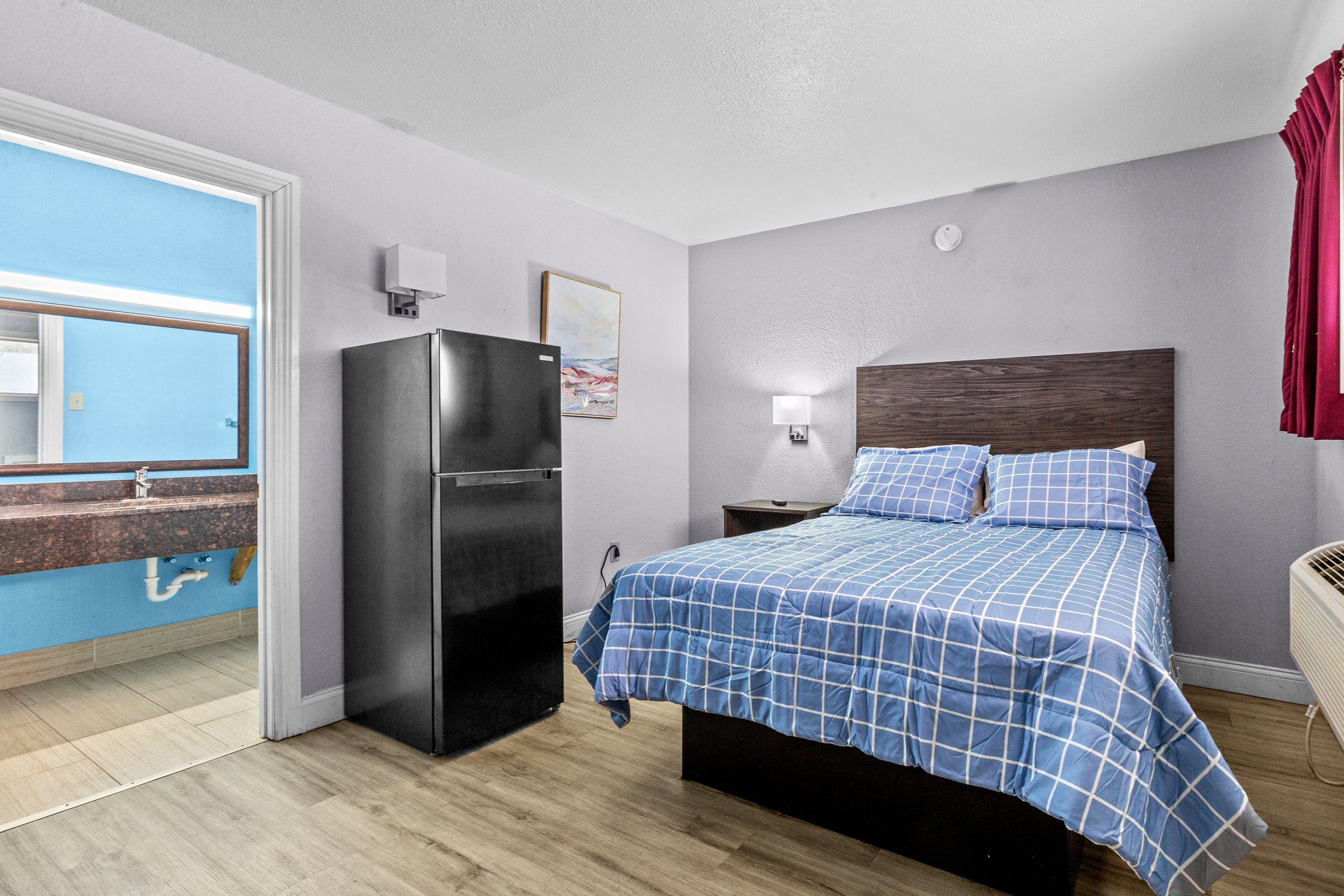 A hotel room with a bed covered in blue checkered bedding, a wooden headboard, a nightstand with a lamp, and a painting on the wall. There is a small refrigerator next to the bed and a window with red curtains. An open doorway reveals a bathroom area with a sink and blue walls.