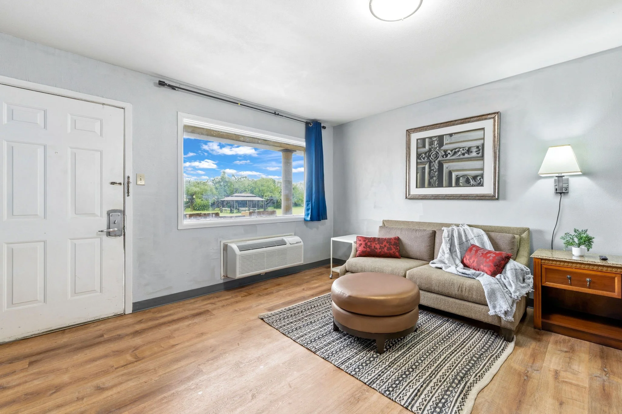 Living room with a beige sofa, red pillows, white blanket, small side table, table lamp, large window with blue curtain showing trees and blue sky, framed artwork, wooden flooring, and front door.