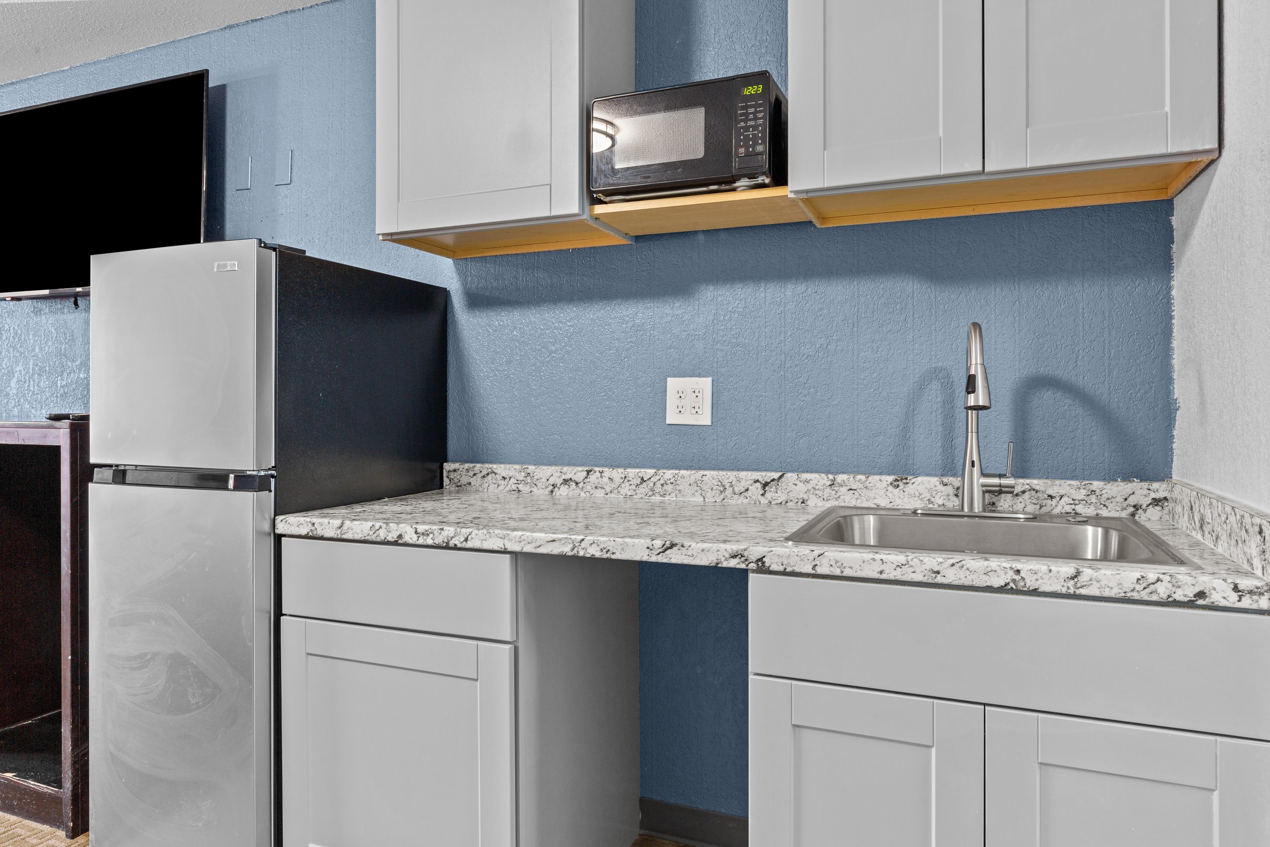 A kitchen area with light gray cabinets, a speckled white and gray granite countertop, a stainless steel sink with a modern faucet, a black microwave on a wooden shelf, a silver and black refrigerator, and a blue painted wall with a white electrical 