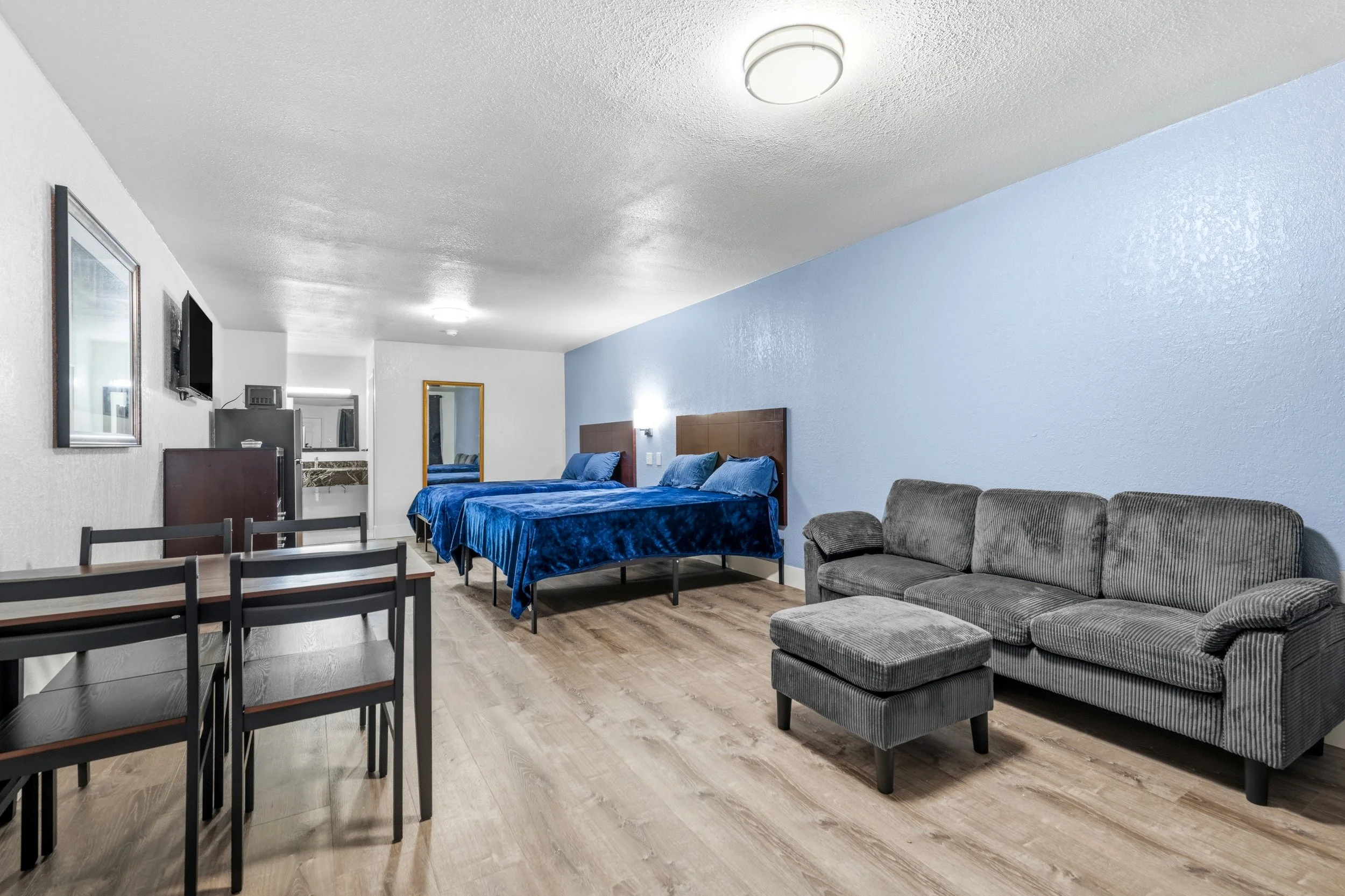 Interior of a hotel suite with two beds with blue bedding, a gray sofa with matching ottoman, a dining table with chairs, a wall-mounted TV, and a mirror.