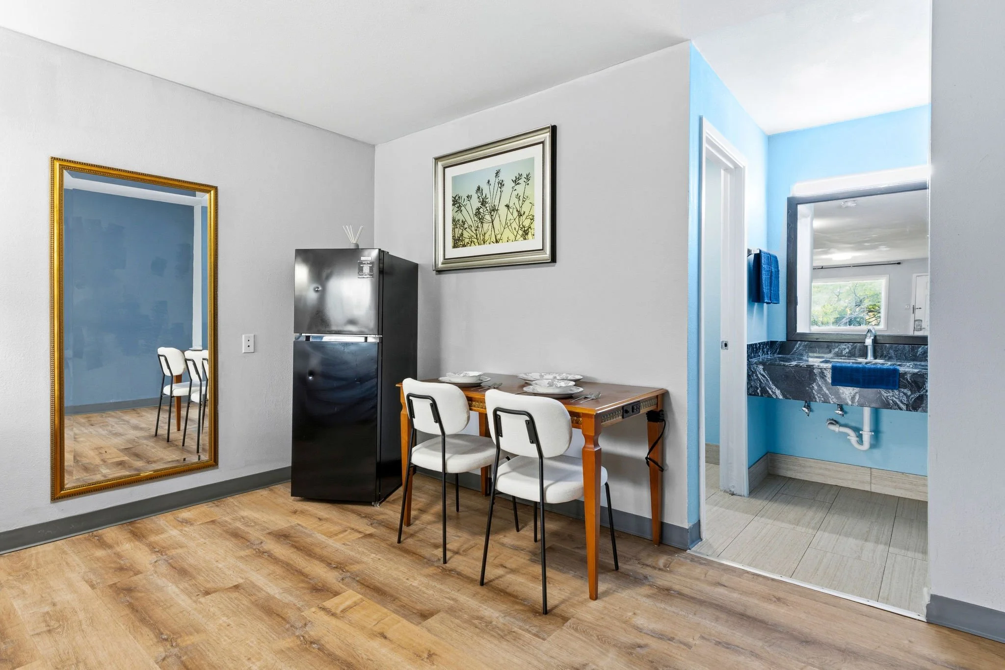 Interior of a home showing a dining area with a wooden table set with white plates and four white chairs, a large mirror with a gold frame on the wall, a framed botanical print, and a visible part of a kitchen with a stainless steel refrigerator and 