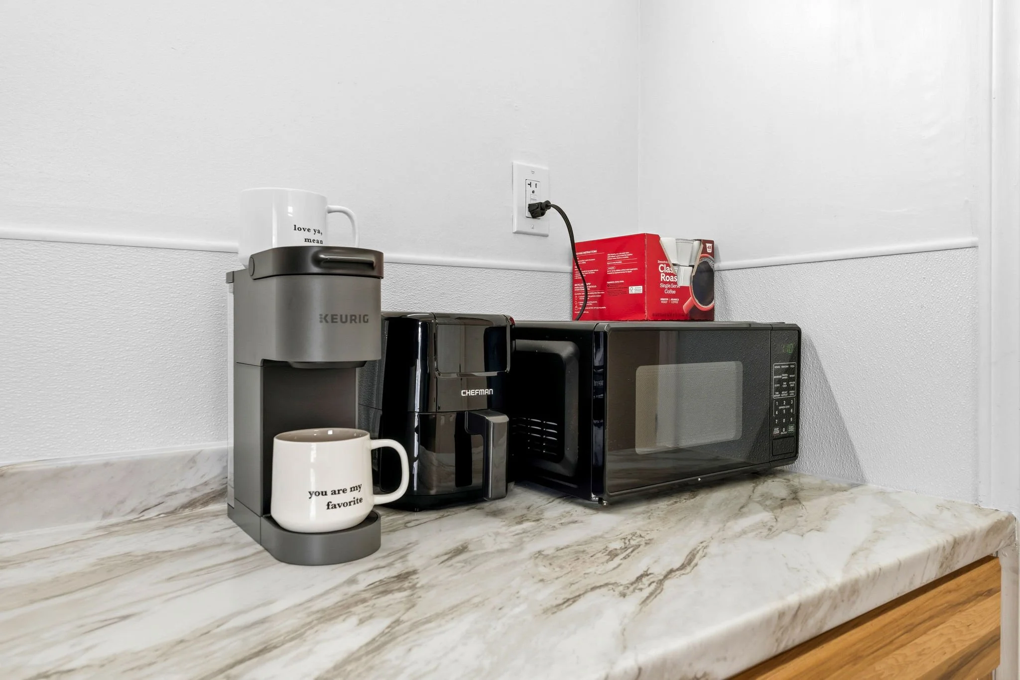 A kitchen countertop with a Keurig coffee maker, a white mug with text, a microwave, and a box of coffee filters against a white wall with an electrical outlet.