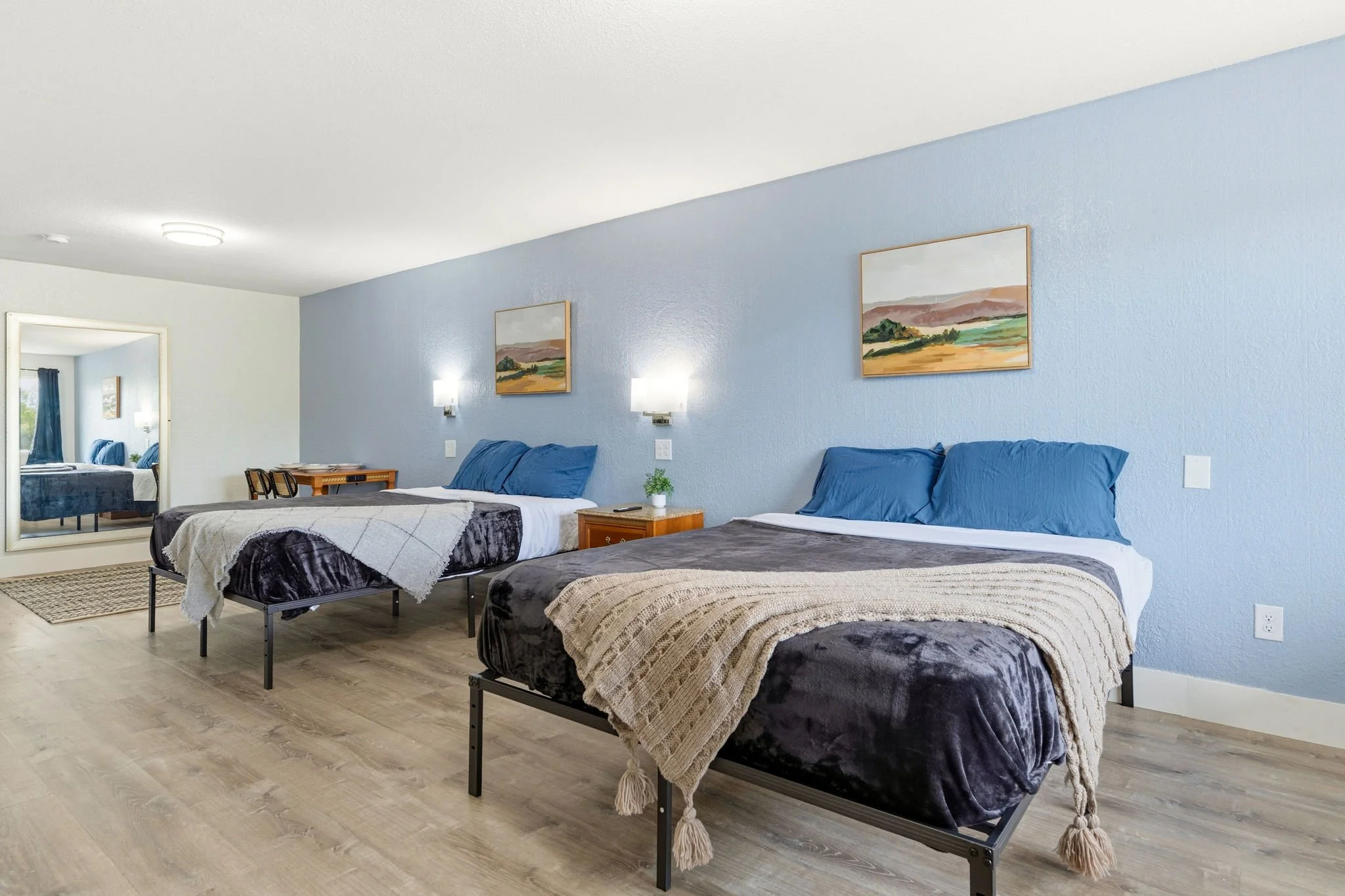 A bright hotel room with three beds, each with blue pillows and blankets. The wall behind the beds is painted light blue and decorated with two landscape paintings. There are wall-mounted lights above each bed, and a bedside table with a small plant.