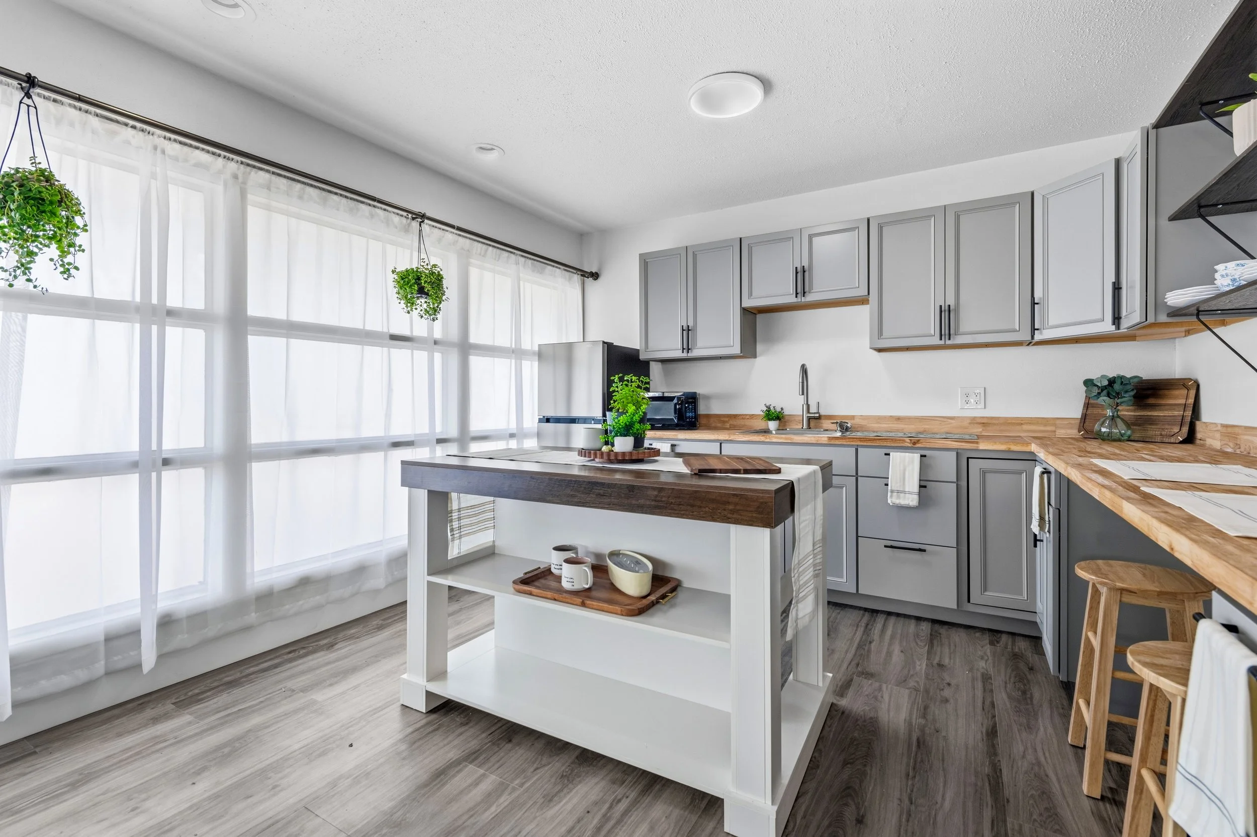 A modern kitchen with gray cabinets, wooden countertops, and large windows with white curtains, decorated with hanging plants and small potted plants on the counter and kitchen island.