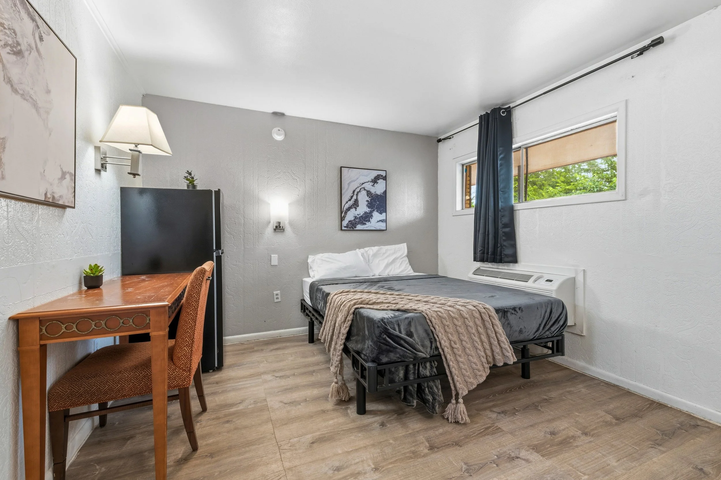 A small bedroom with a single bed with dark gray bedding and a beige blanket, a wooden desk with a patterned chair, two abstract art pieces on white walls, two windows with black curtains and a view of green trees outside, and a wooden floor.