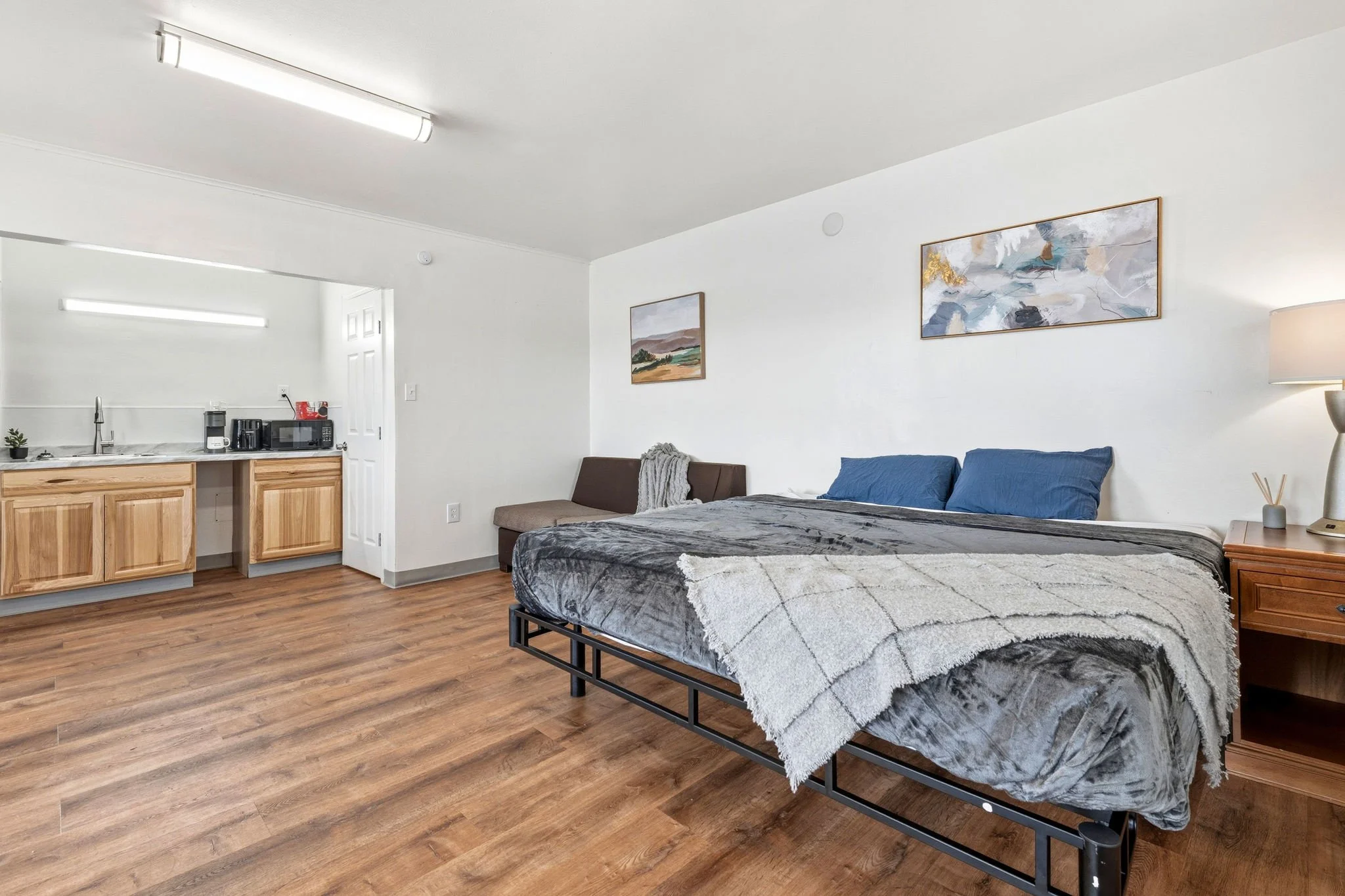A bedroom with a bed covered in dark gray and white bedding, a wooden nightstand, wall art, and a small sitting area with a bench and blanket. The room has white walls and wood flooring.