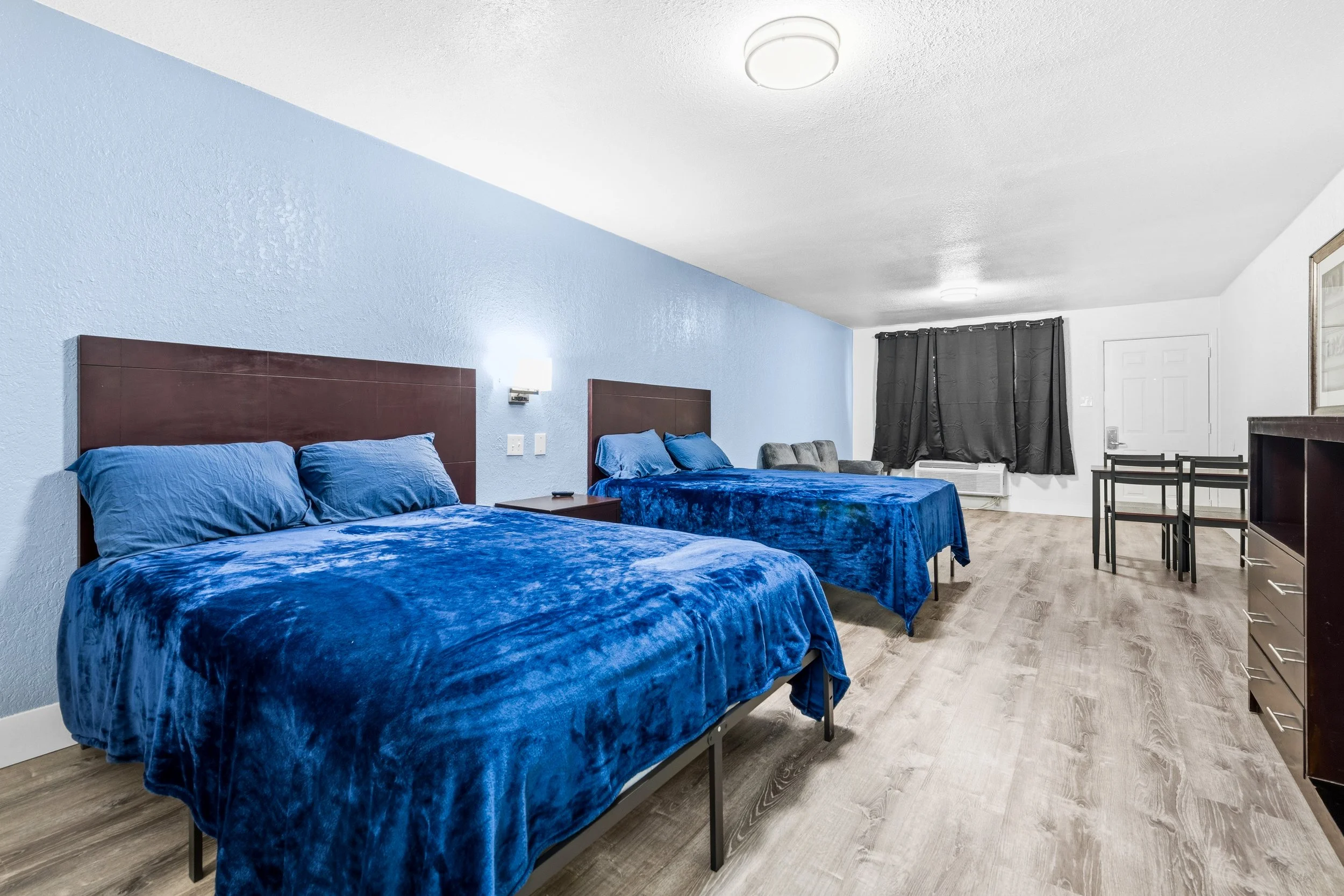 Two beds with blue velvet bedspreads and matching pillows, headboards, a chair, a window with black curtains, and a dresser in a hotel room with wood flooring.
