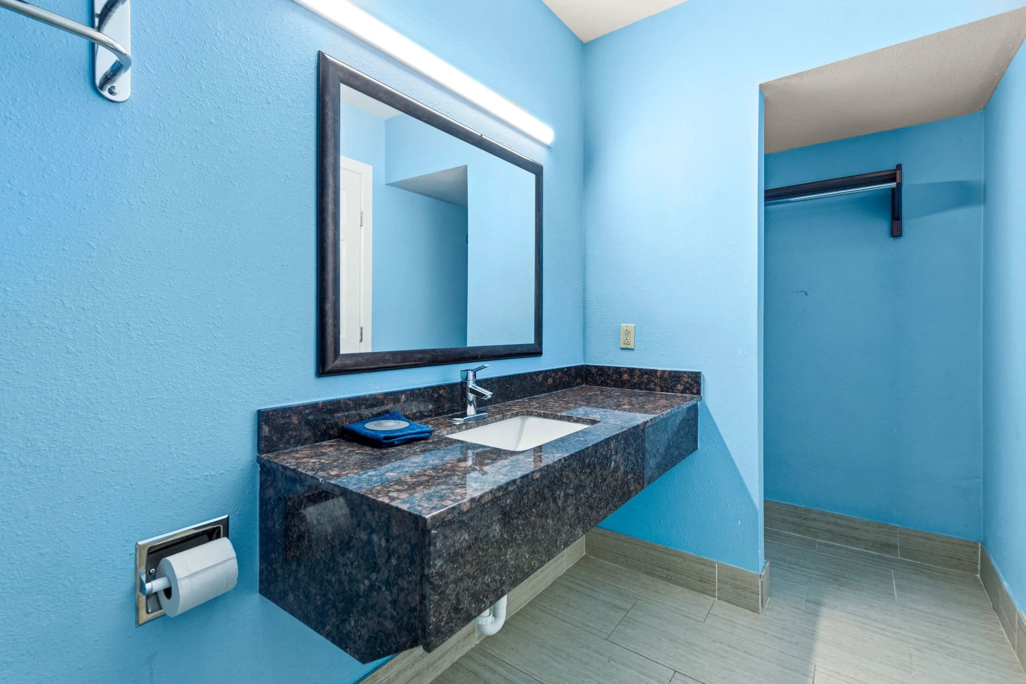 A bathroom with light blue walls, a rectangular mirror with a dark frame, and a granite countertop with a white rectangular sink. There is a faucet, a tissue box, and a blue item on the counter. A wall-mounted reflector and a toilet paper roll are vi