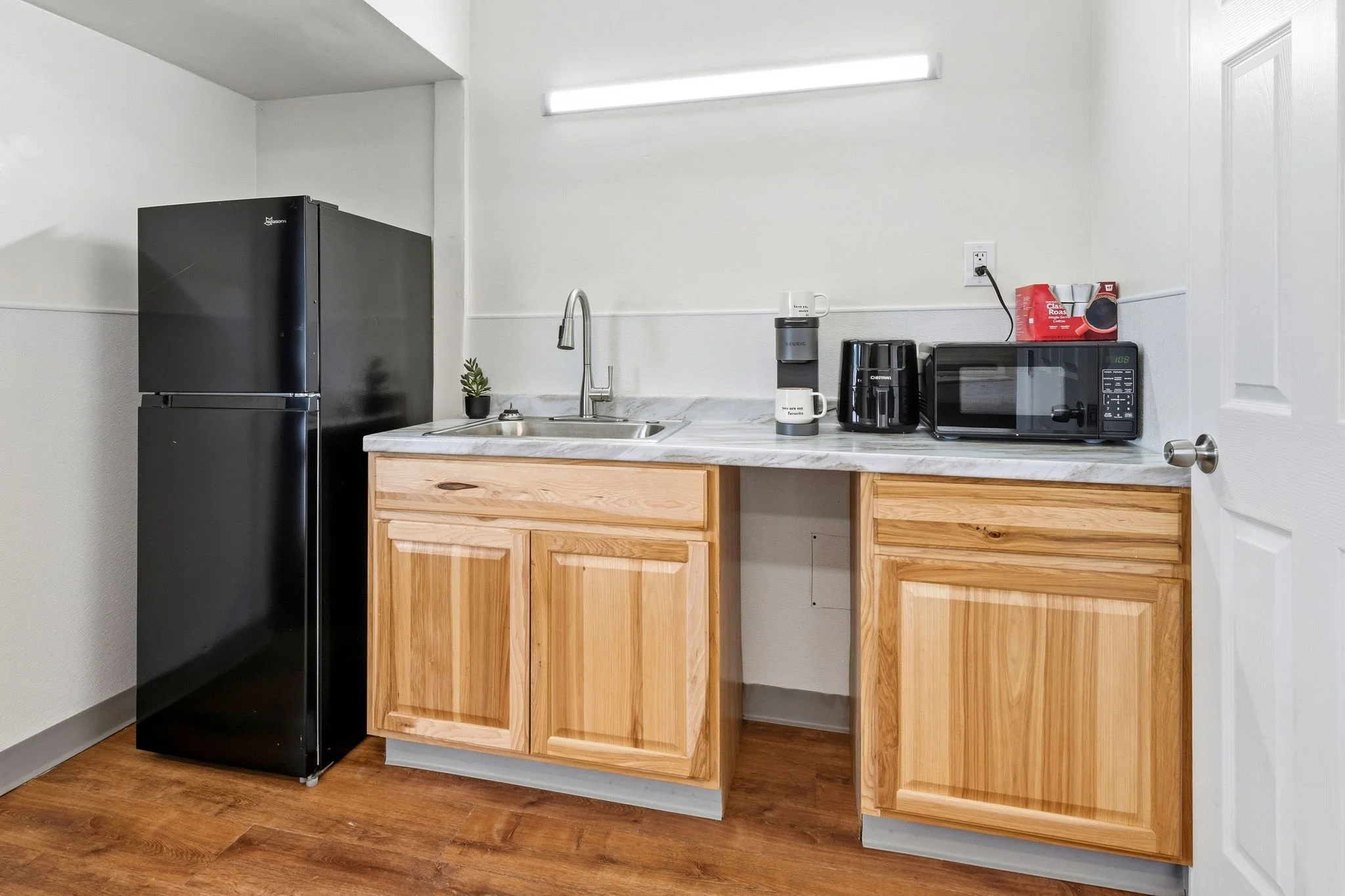 A kitchen with light wood cabinets, a black refrigerator, a small sink, a coffee maker, a microwave, and red packaging on top of the microwave against a white wall with a fluorescent light fixture.