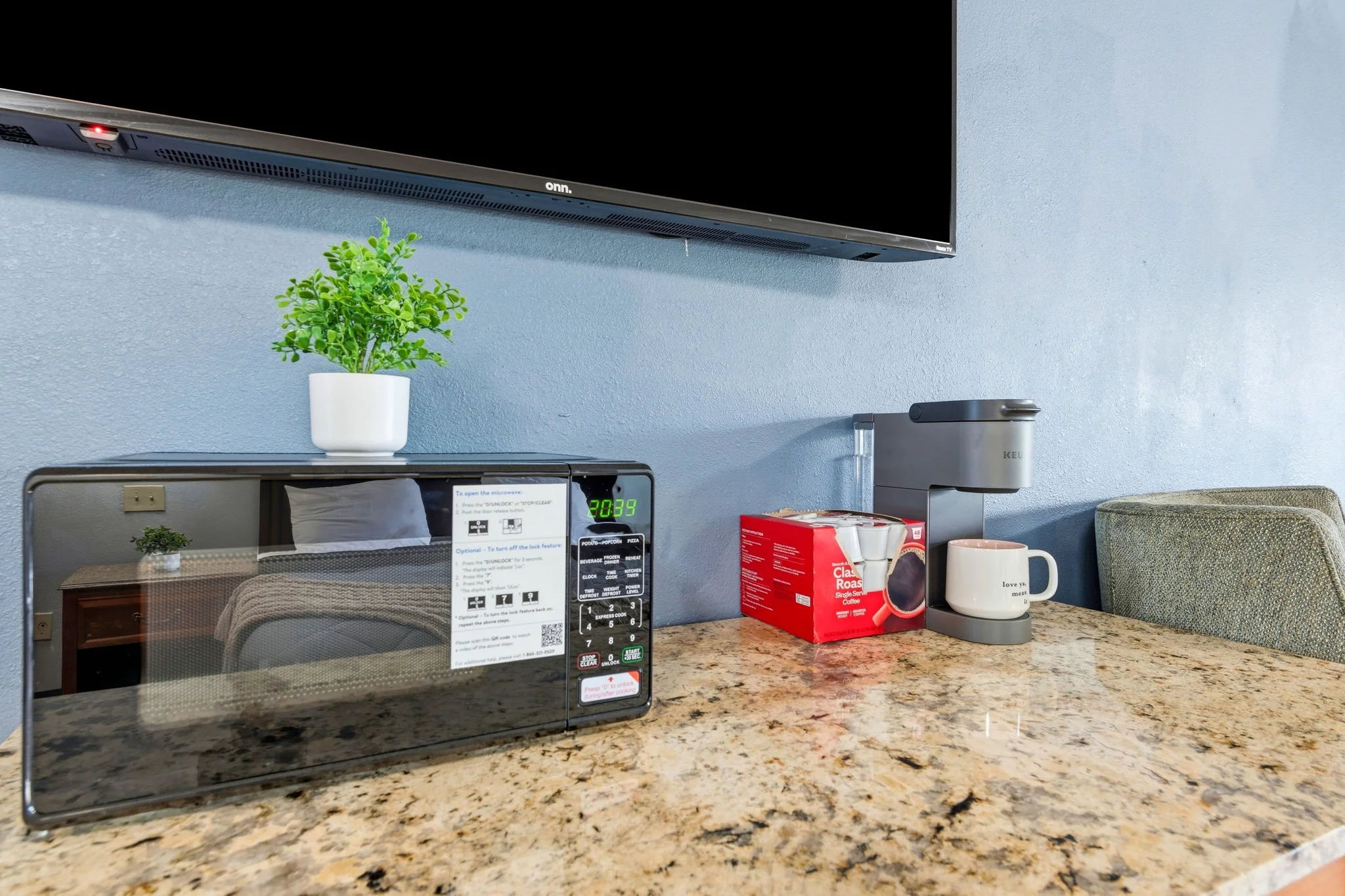 A kitchen countertop with a microwave, a green potted plant, a red box of coffee, a white mug, and a coffeemaker; a television mounted on the wall above.
