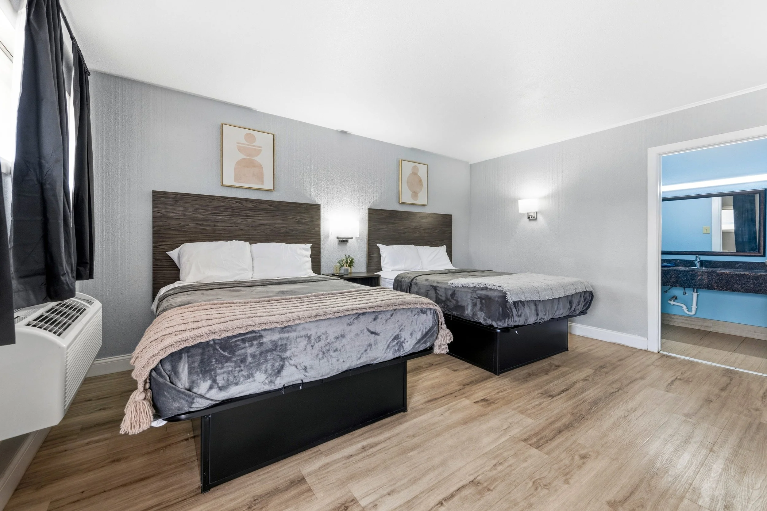 Two beds with dark wood headboards and gray bedding in a hotel room, with abstract art on the wall above and a window with dark curtains to the left.