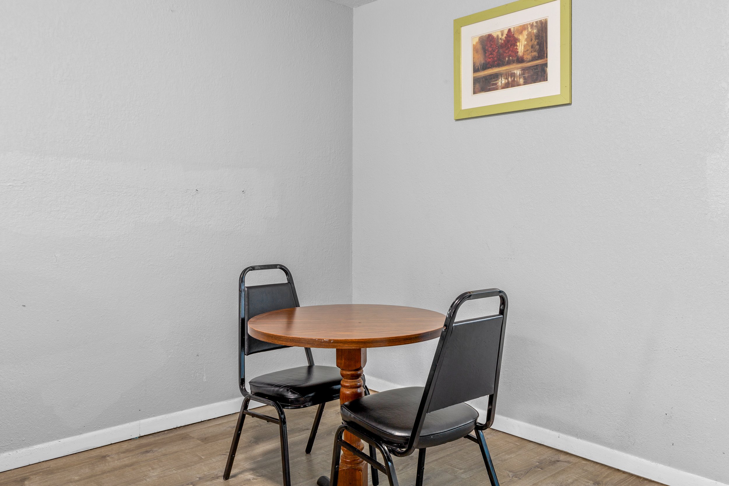 A corner of a room with a small round wooden table and three black chairs. There is a painting of trees with autumn foliage on the wall.