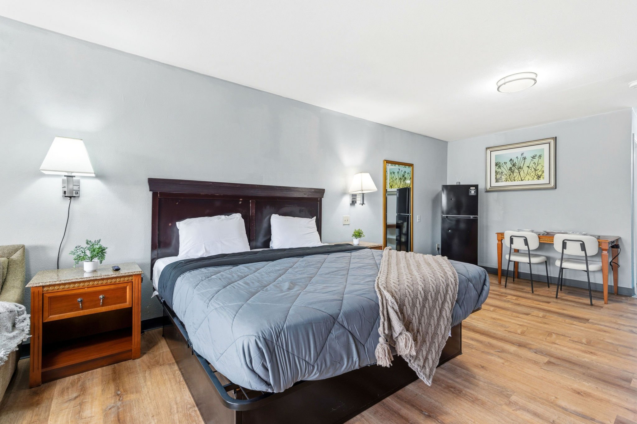 Interior of a hotel room with a bed, two wall-mounted lamps, a small nightstand with a plant, a desk with two chairs, and framed nature artwork on the walls.