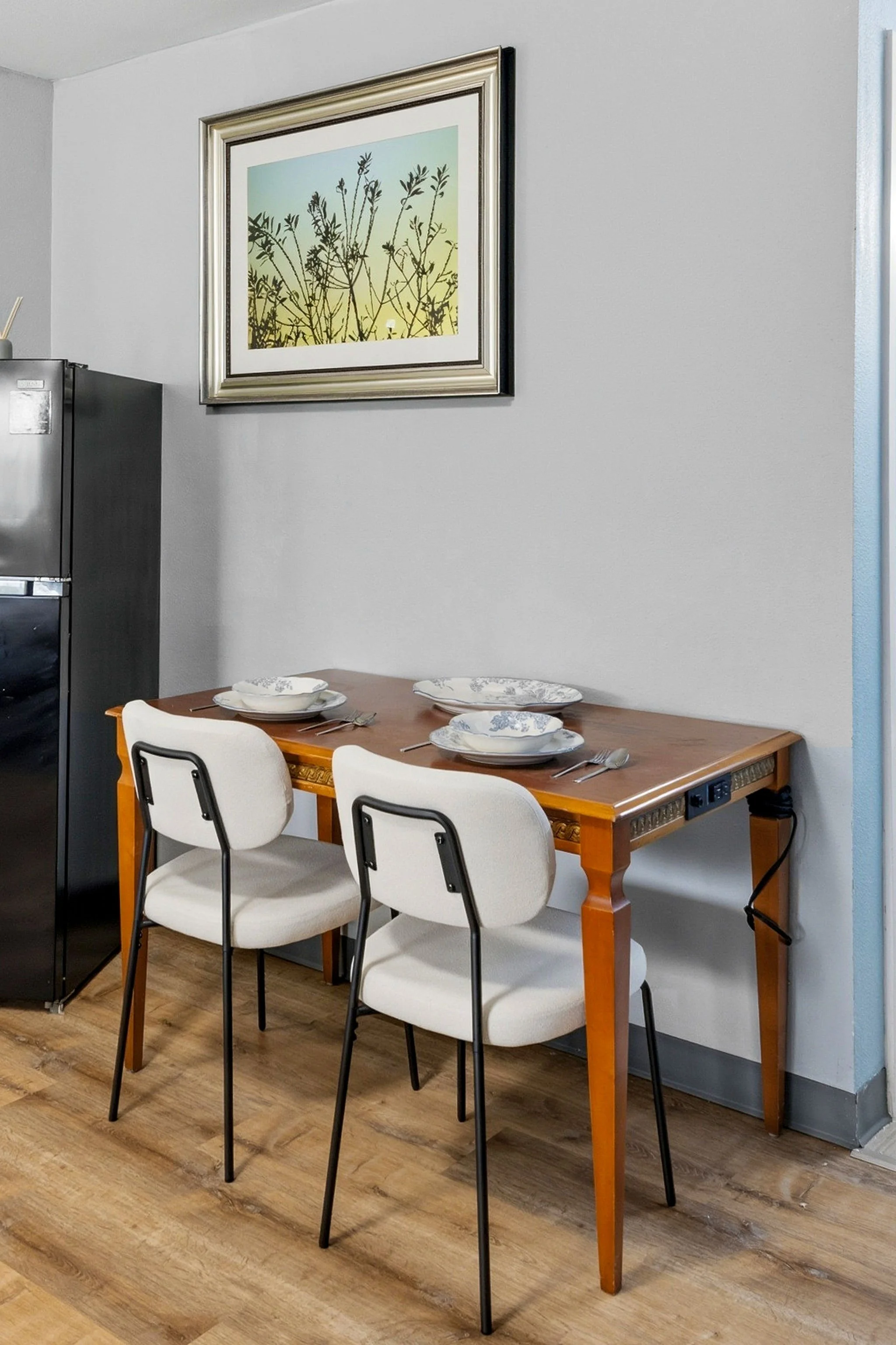 A dining area with a wooden table set for three, with three white chairs. The table has plates, bowls, and utensils arranged for a meal. Above the table, there is a framed artwork of silhouetted plants on a gradient background. To the left, part of a