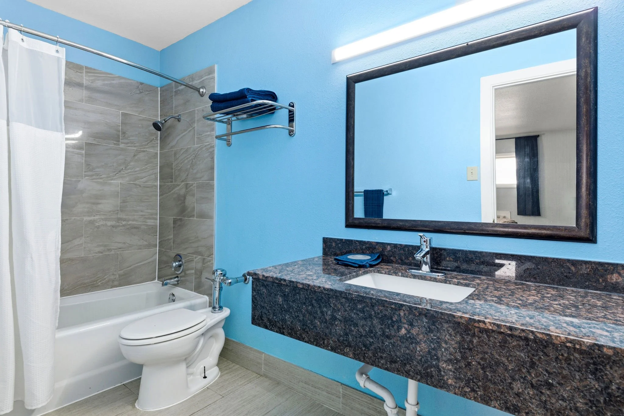 A small bathroom with a bathtub and shower on the left, a toilet in front of the bathtub, and a vanity with a sink on the right. The bathroom has light blue walls, a large mirror, a granite countertop, and towels on a shelf and towel bar.