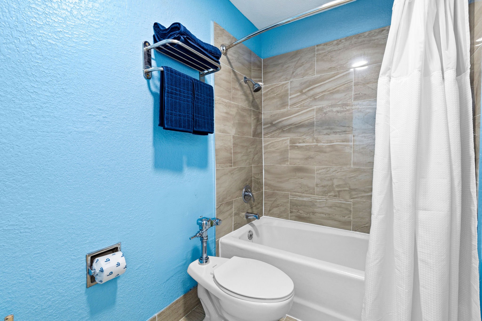 A bathroom with a blue textured wall, a white bathtub with a shower curtain, a monitor above the toilet paper holder, and a black towel hanging on a silver towel rack on the blue wall.