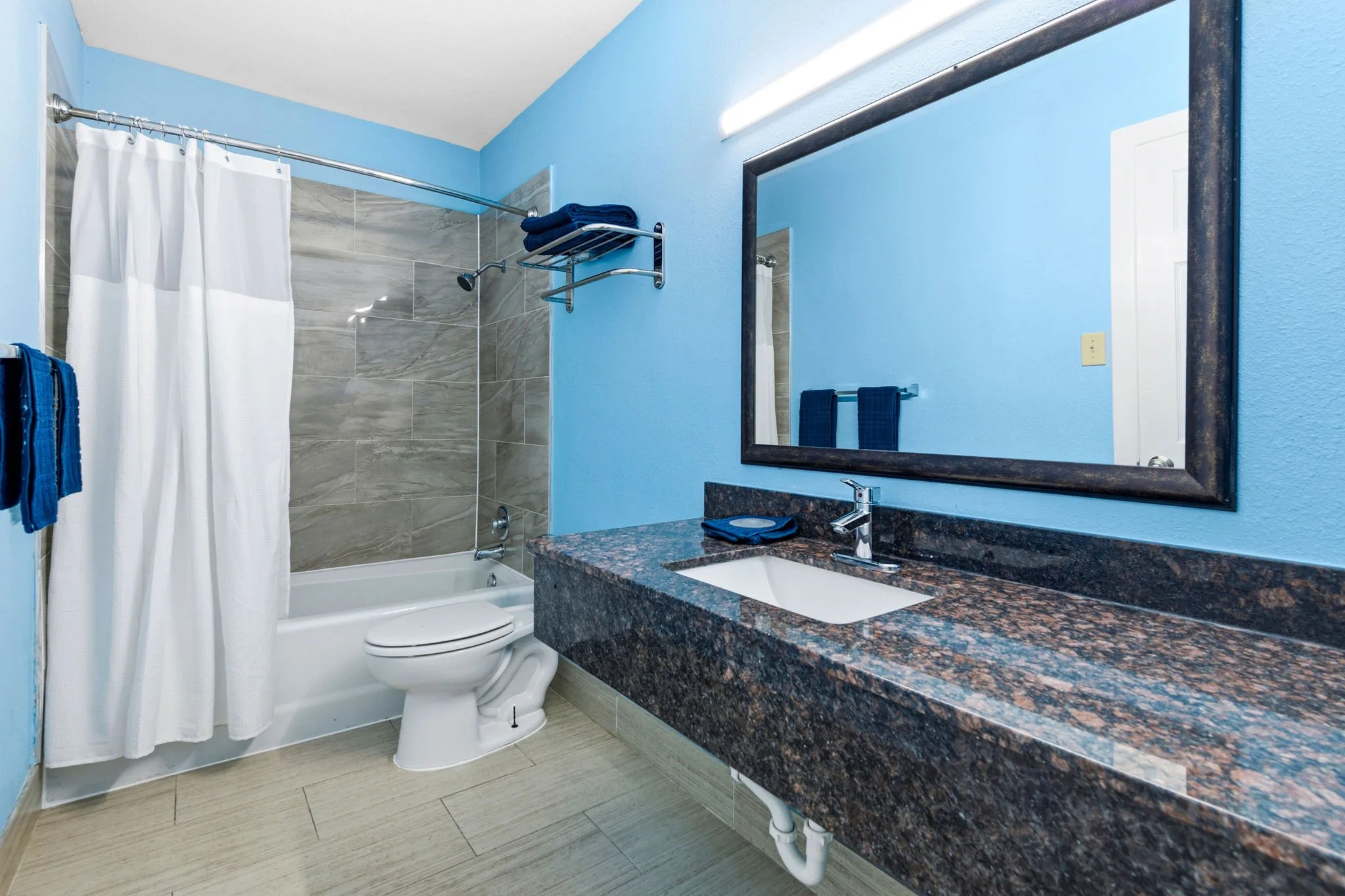 Bathroom with blue walls, a granite countertop with a white sink, large mirror, bathroom towel rack, and a shower with a white curtain and gray tiled walls.