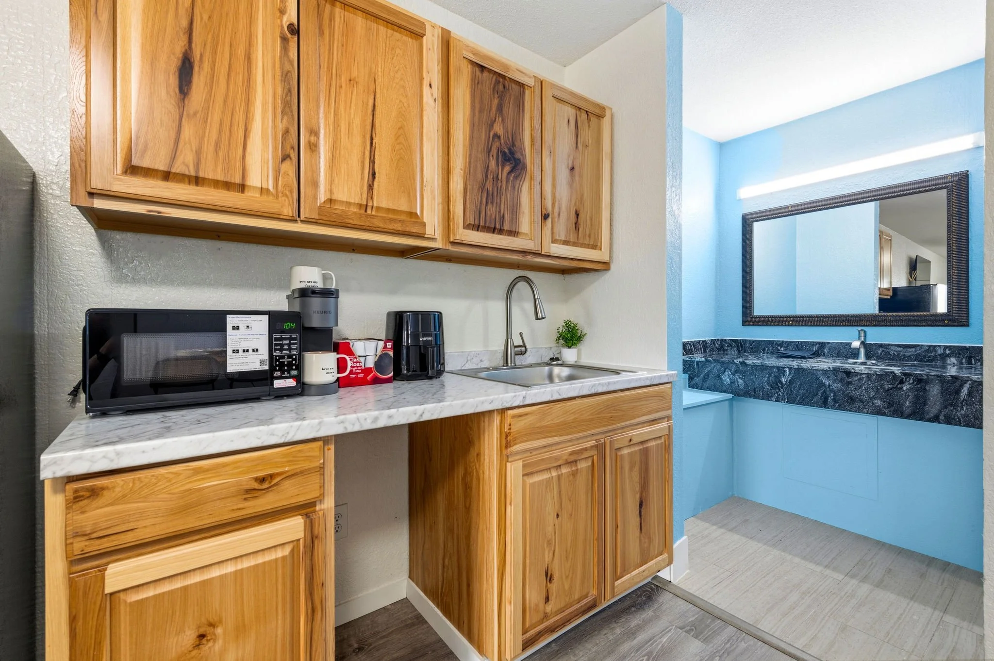 Kitchen area with wooden cabinets, a microwave, coffee maker, and a small plant on a marble countertop. There is a mirror on the wall reflecting part of the room, and the wall behind is painted light blue.