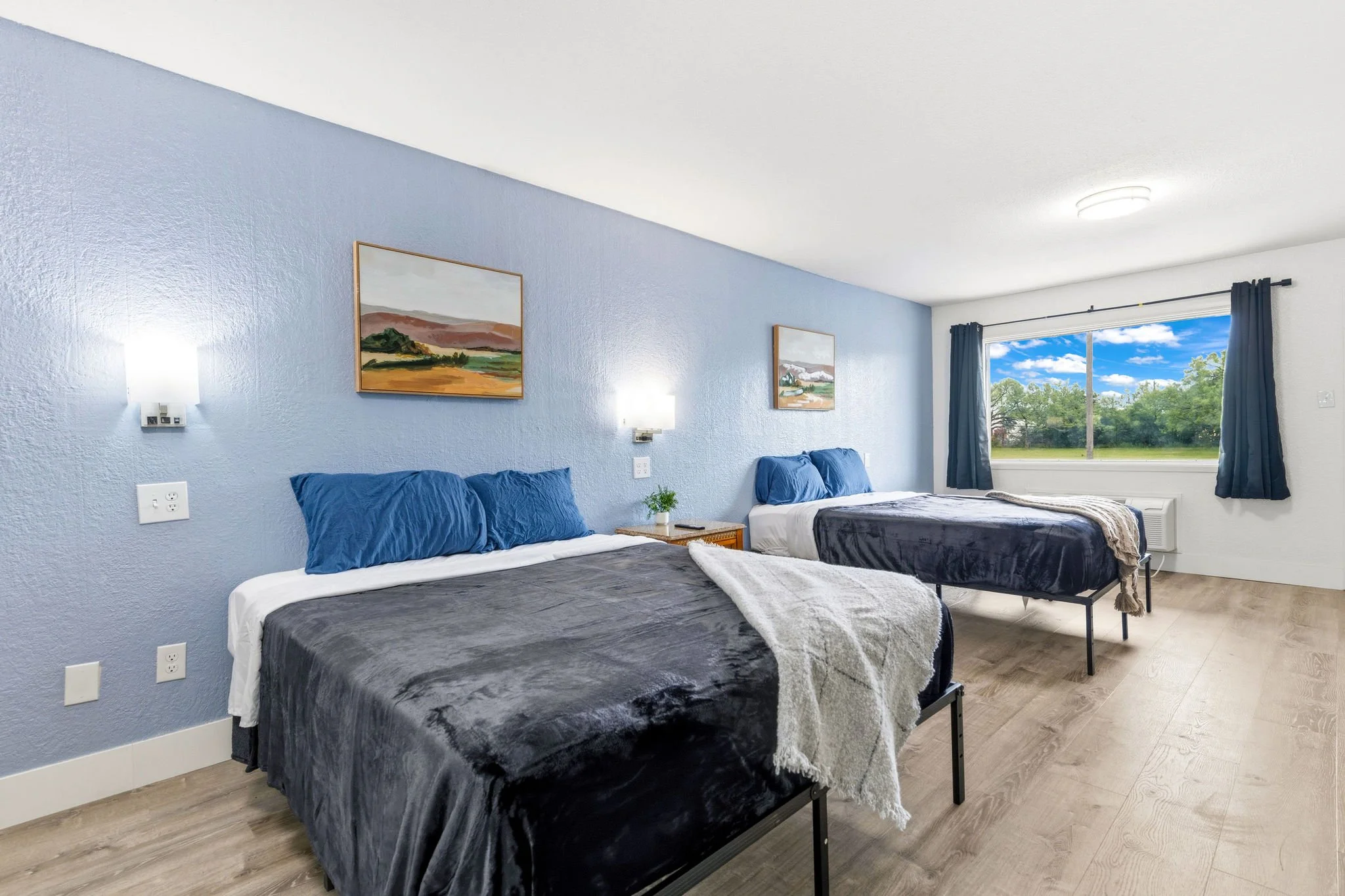 A hotel room with two beds, blue and gray bedding, artwork on the light blue wall, and a large window with blue curtains overlooking a green landscape with trees and a blue sky.