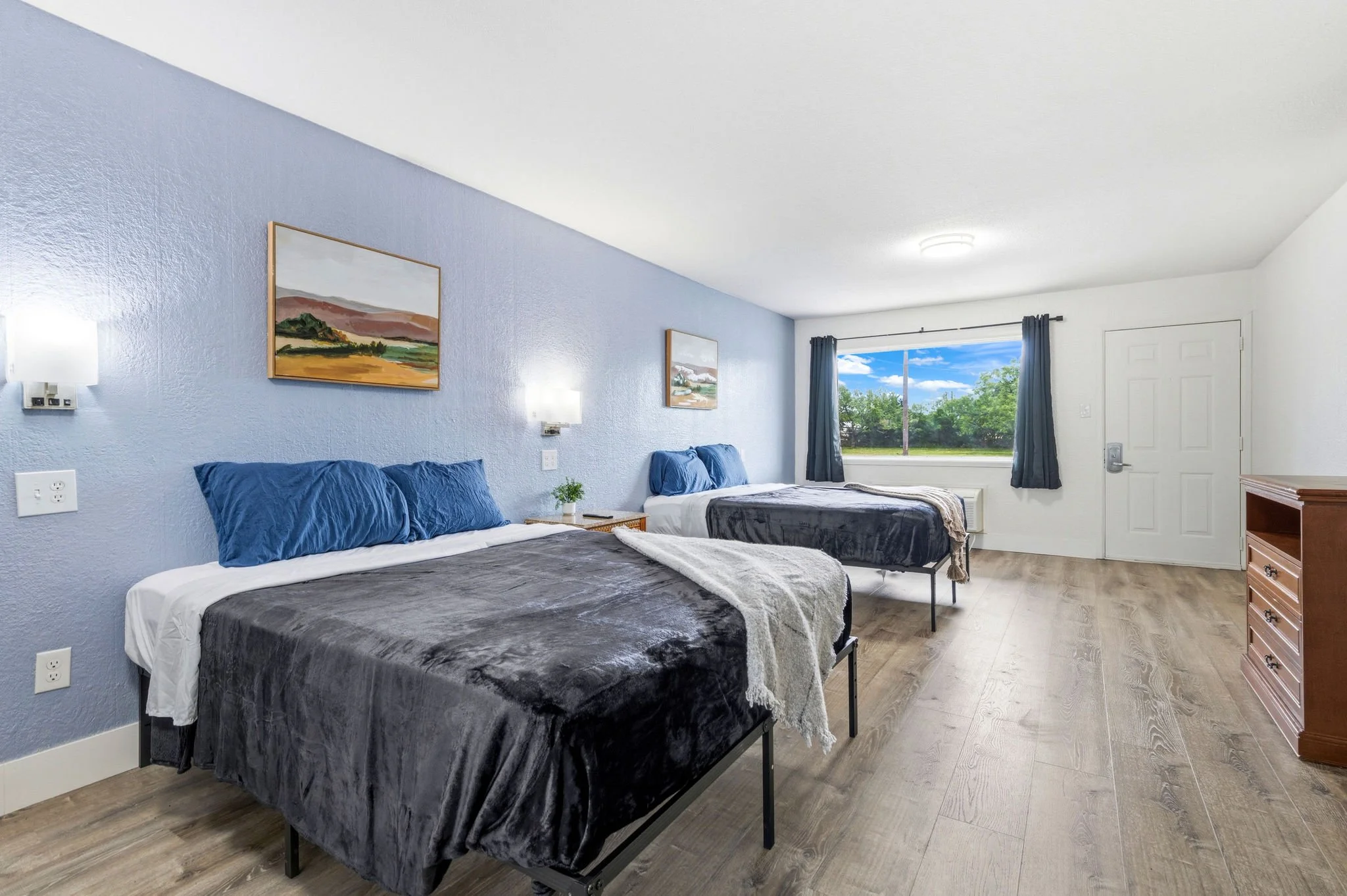 A bright hotel room with two beds, each with a dark gray comforter and blue pillows, a wooden nightstand with a plant, artwork on the blue accent wall, a large window showing trees and a blue sky, and a white door.