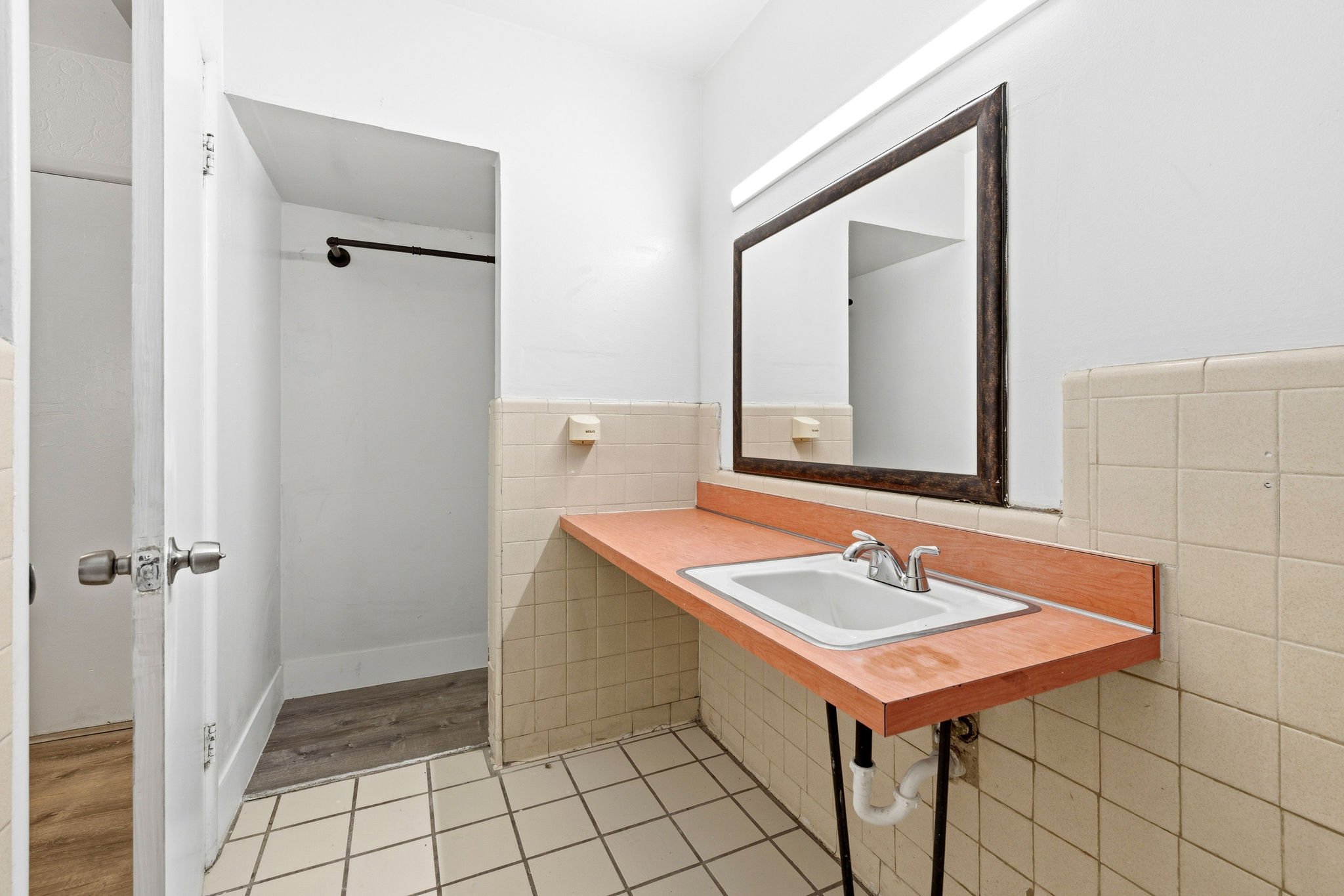Small bathroom with white walls, a rectangular vanity with a white sink and silver faucet, a large mirror with a rusted frame, beige tile backsplash, beige tile walls, white tiled floor with black grout, a paper towel dispenser, and a white door with