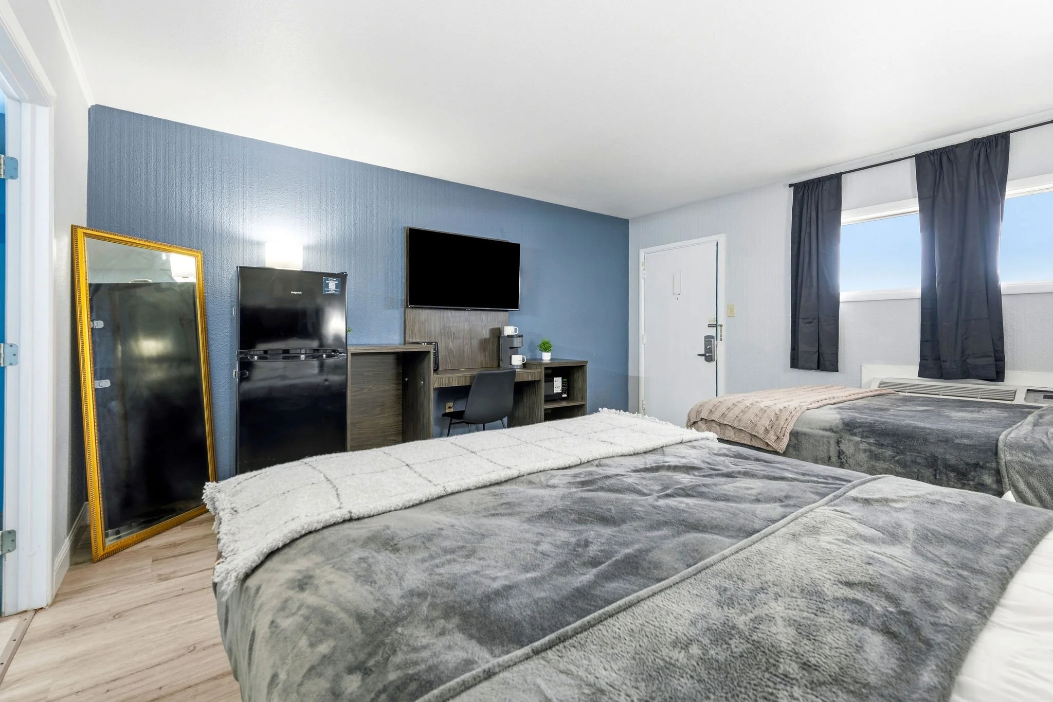 A hotel room with two beds covered in gray blankets and beige throws. There is a large mirror with a gold frame, a small refrigerator, a flat-screen TV, a desk with a plant, and a black chair. The room has a blue accent wall, white ceiling, and windo