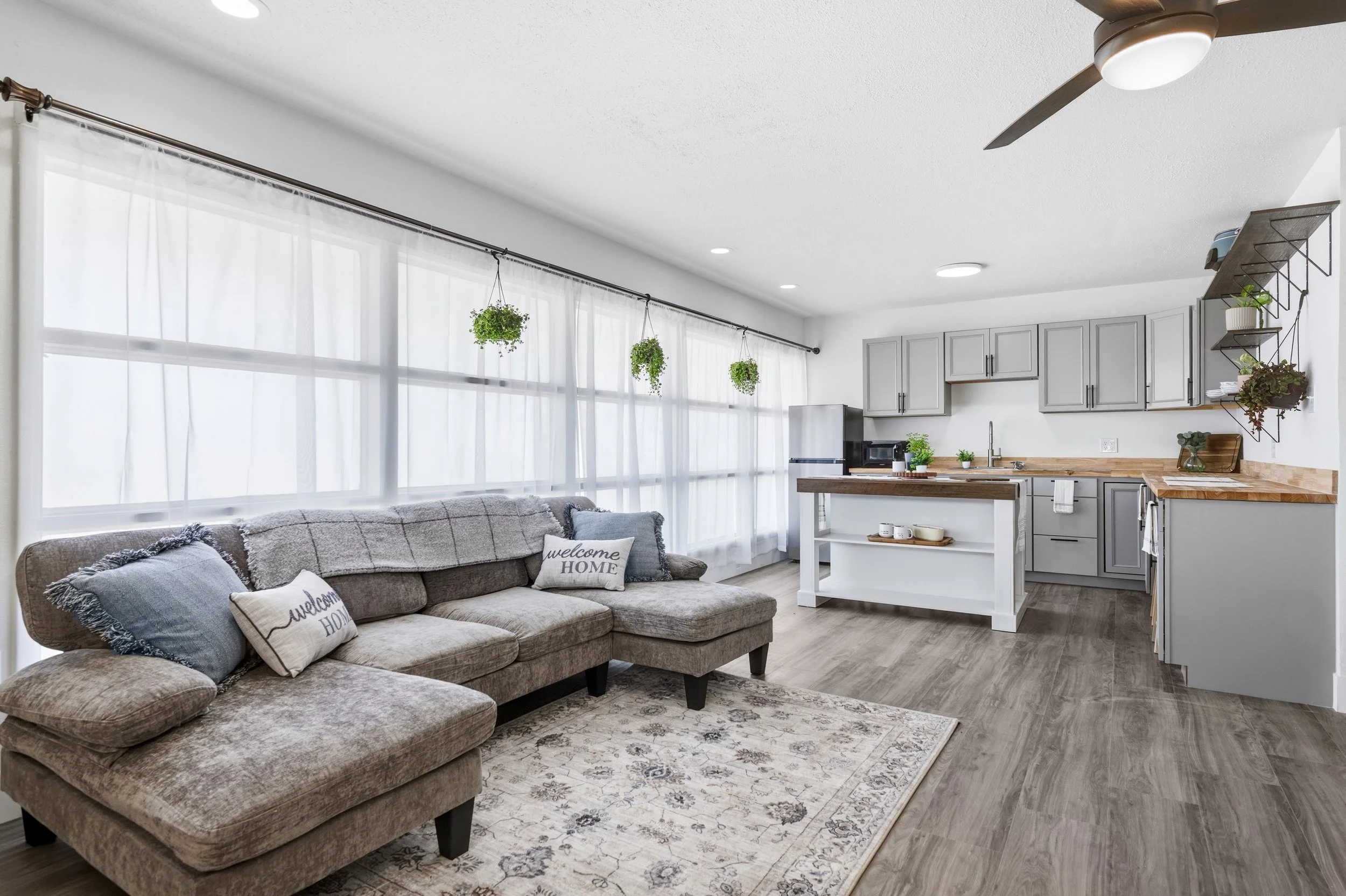 Living room with large window, grey sofa with pillows, area rug, kitchen with grey cabinets, wooden countertops, plants, and hanging planters.
